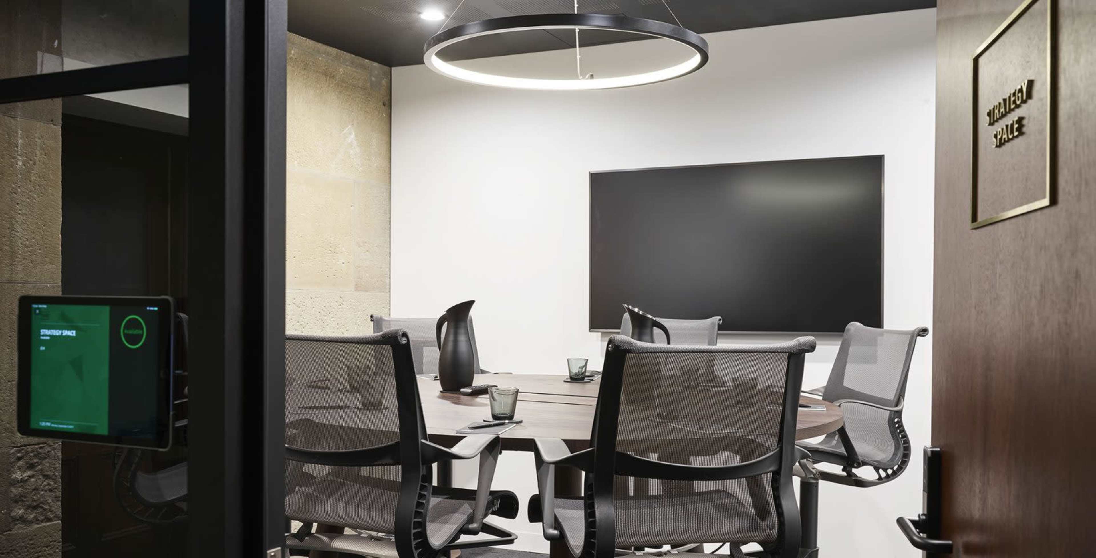 A conference room features a round table surrounded by several mesh chairs, with a large screen mounted on the wall and a door labeled "Meeting Room."