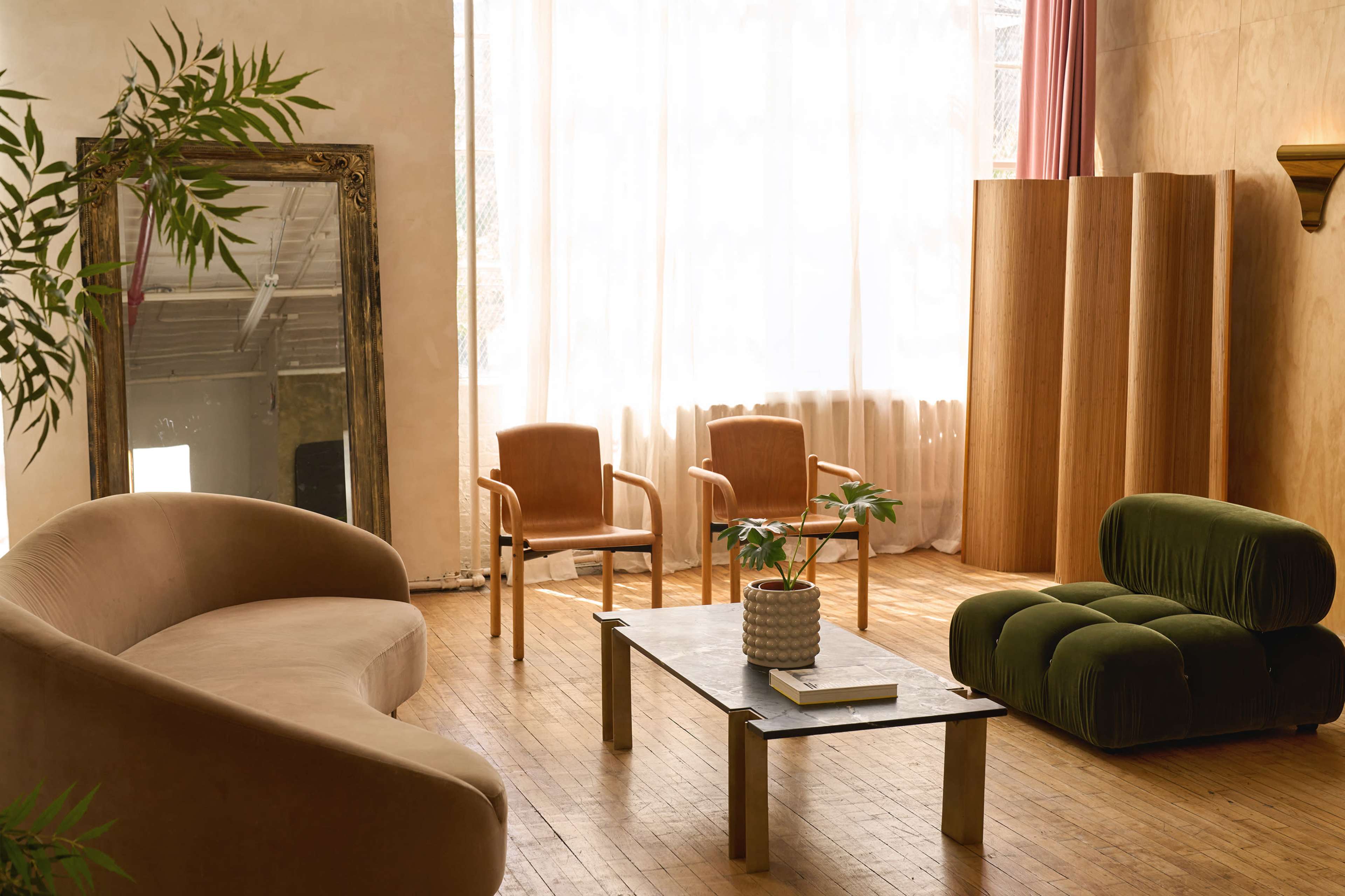The image shows a stylish interior living space featuring a curved beige sofa, two brown chairs, a green velvet chair, a glass coffee table, and a large mirror, with natural light filtering through sheer curtains.