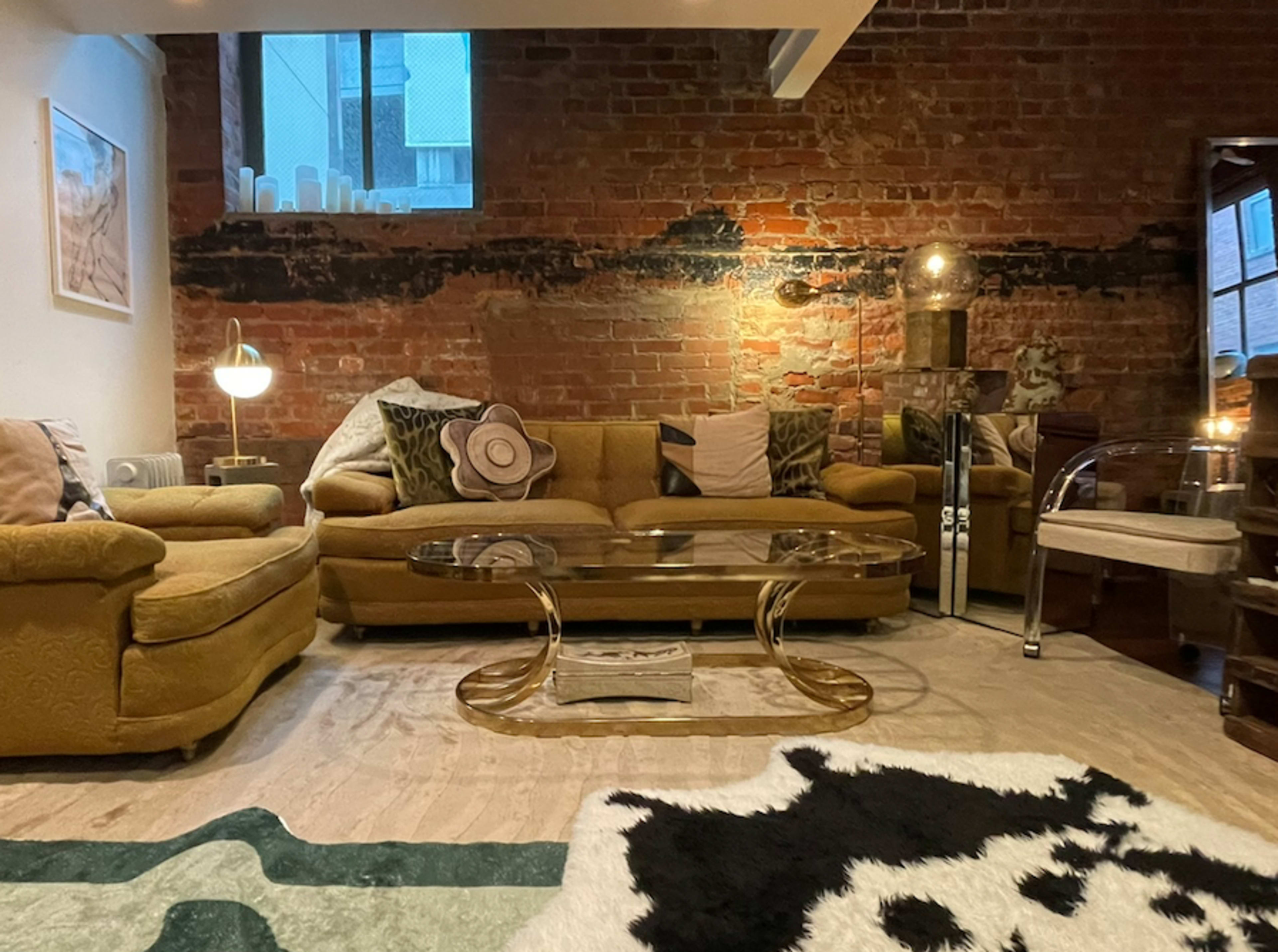 The living area features a pair of yellow sofas, a glass coffee table, and a cowhide rug, complemented by exposed brick walls and various decorative pillows.