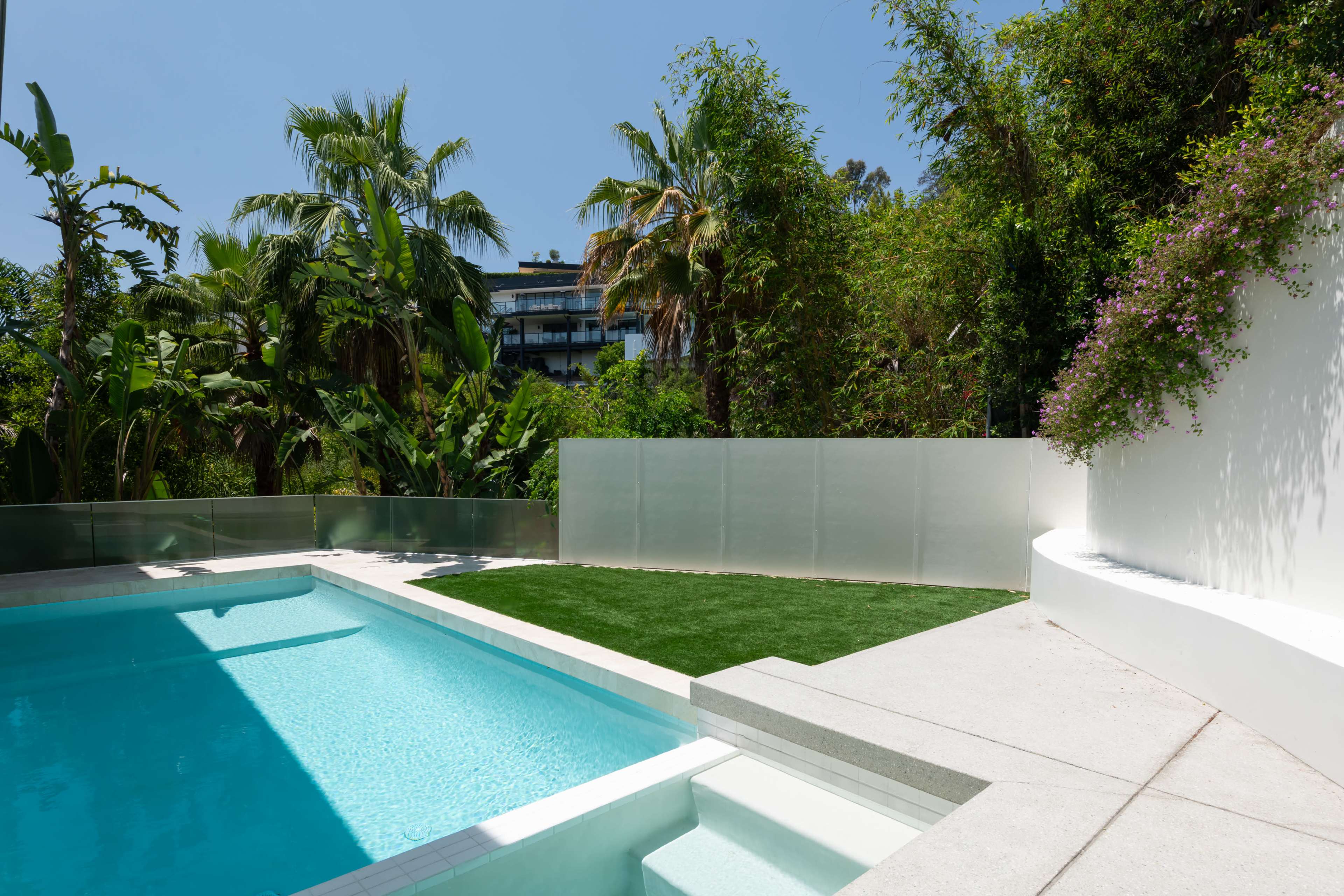 A modern swimming pool is situated next to a landscaped area with artificial grass and surrounded by tropical plants.