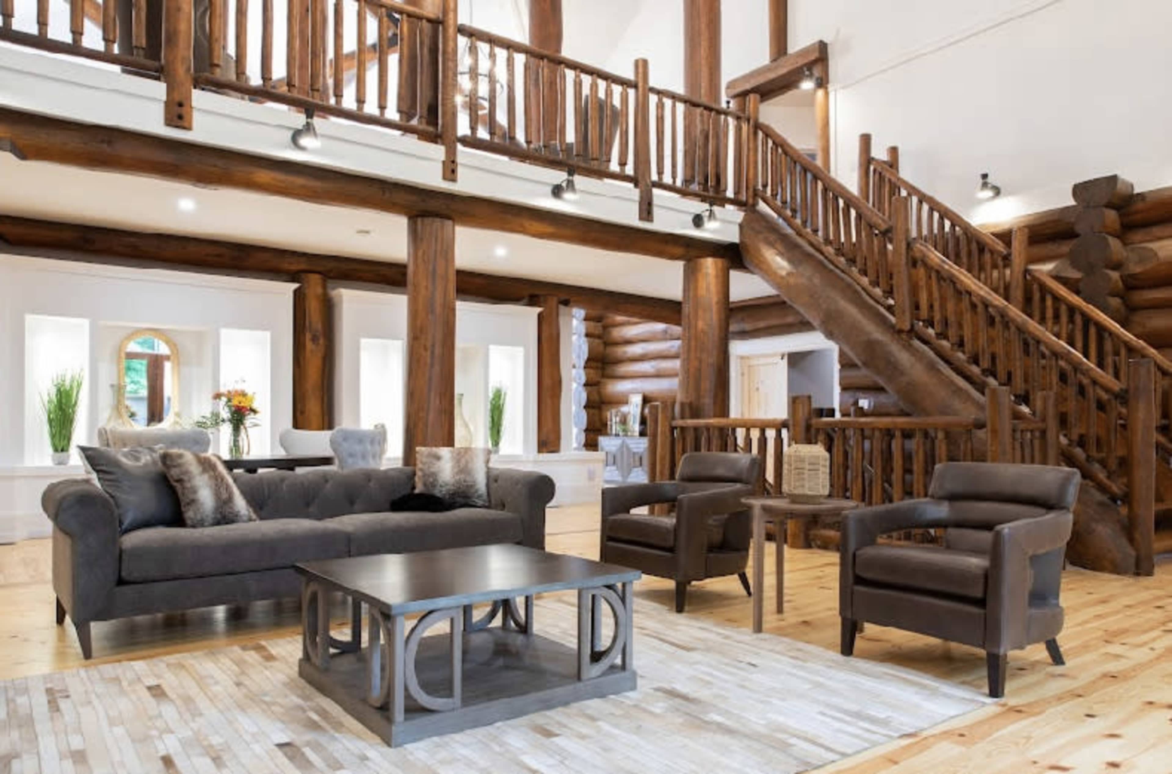 A spacious living room with a wooden log cabin design, featuring a gray sofa, two armchairs, and a central coffee table on a patterned rug, all set beneath a staircase with wooden railings.