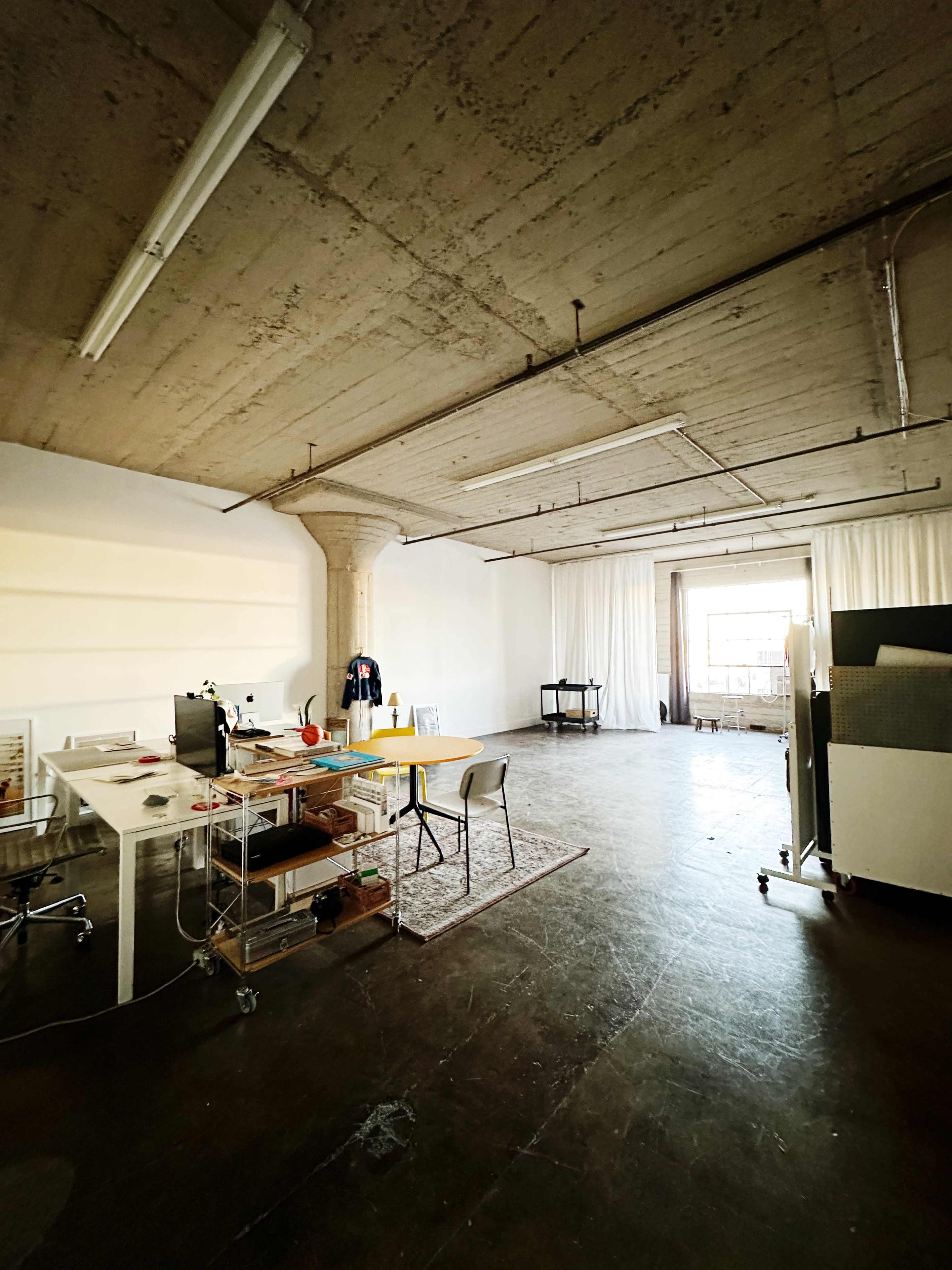 The image shows a spacious, industrial-style office with a large window, a desk with a computer, and minimal furnishings.