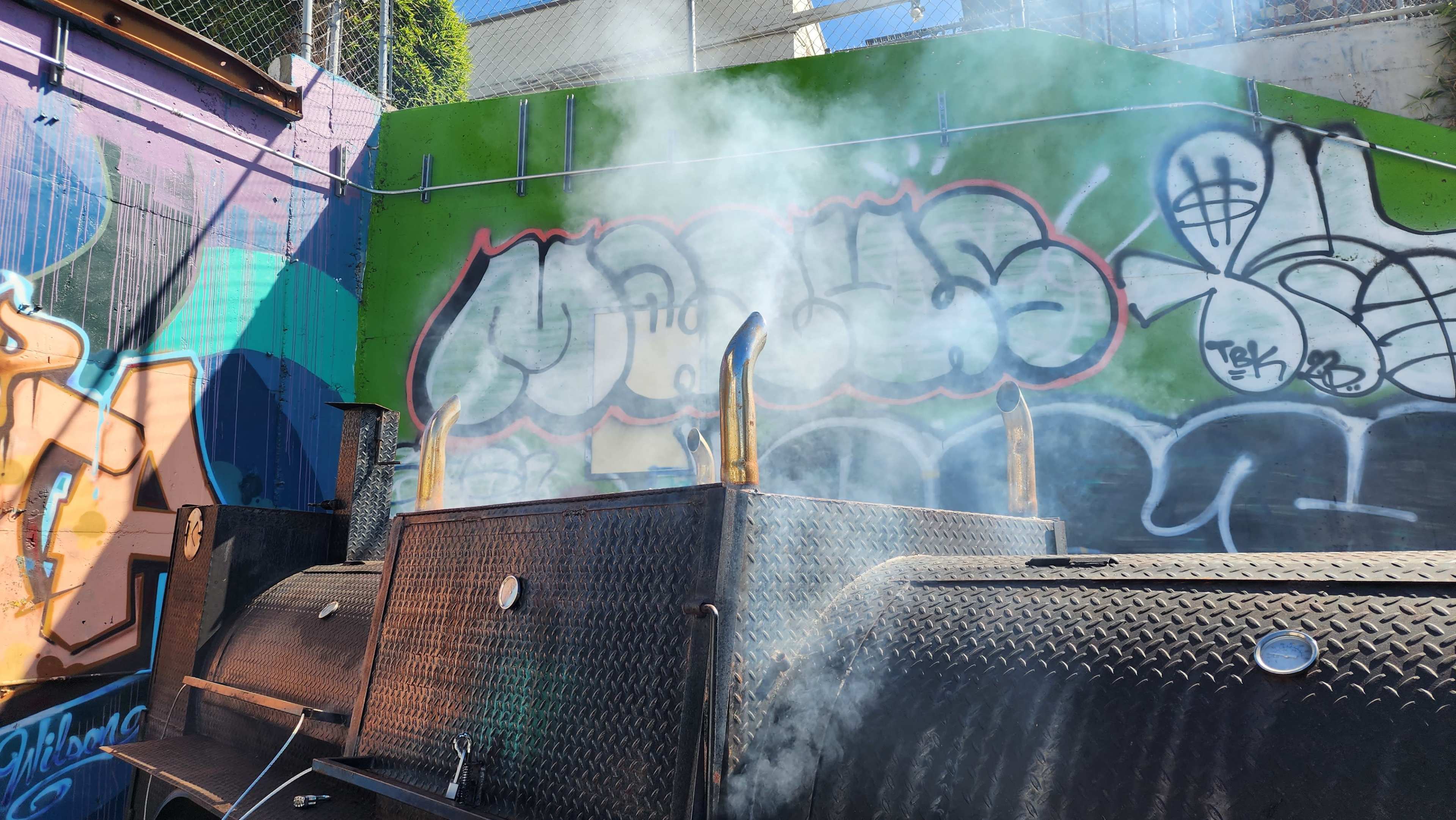 Smoke rises from the chimney of a cooking smoker adjacent to a colorful graffiti-covered wall.