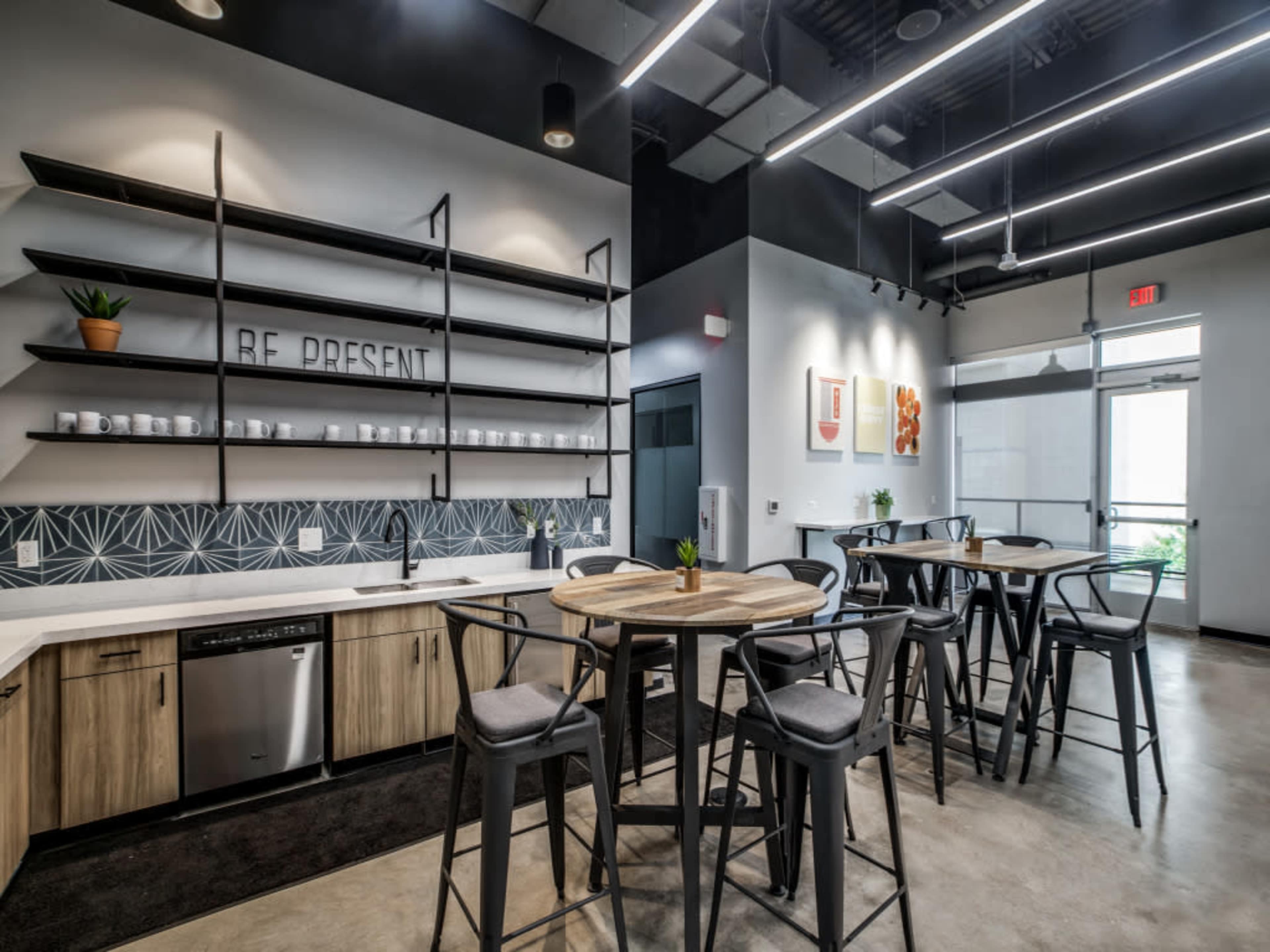 A modern kitchen and dining area with wooden tables, black chairs, open shelving, and large windows letting in natural light.