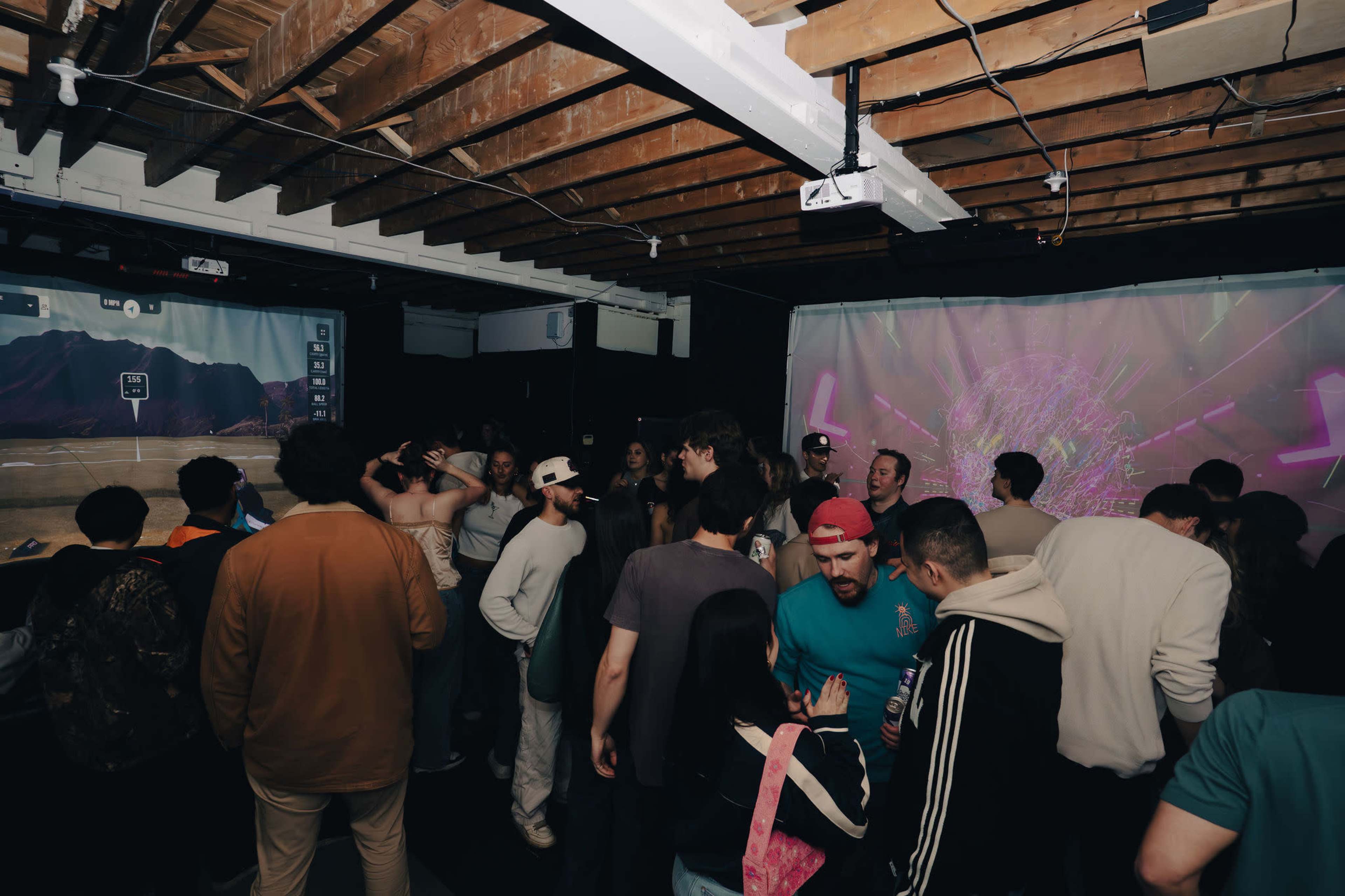 A large group of people interacts and socializes in a dimly lit room with projectors displaying visual graphics on the walls.