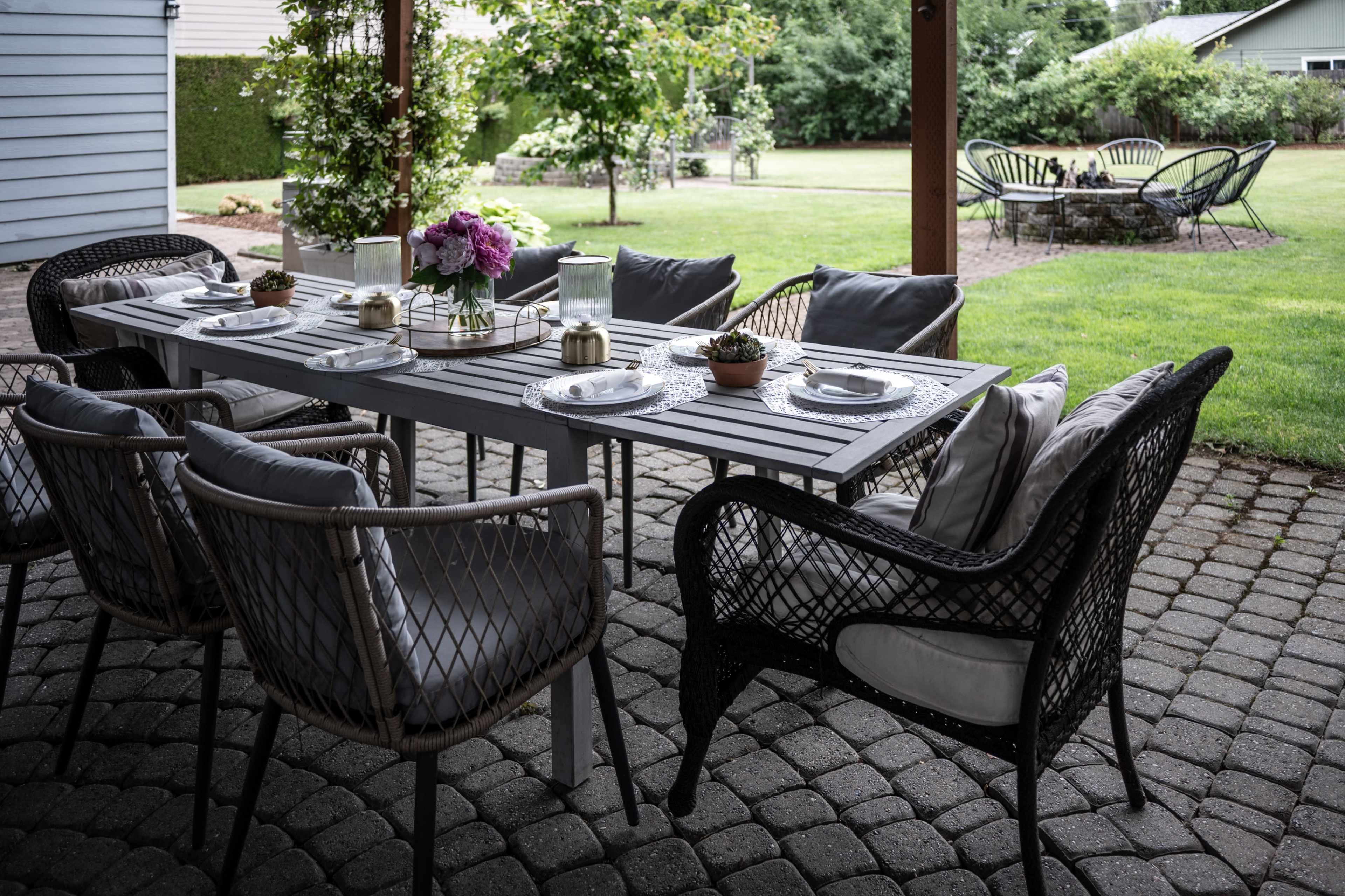 A patio dining table with six chairs is set with dishes and decorated with flowers, surrounded by a landscaped yard.
