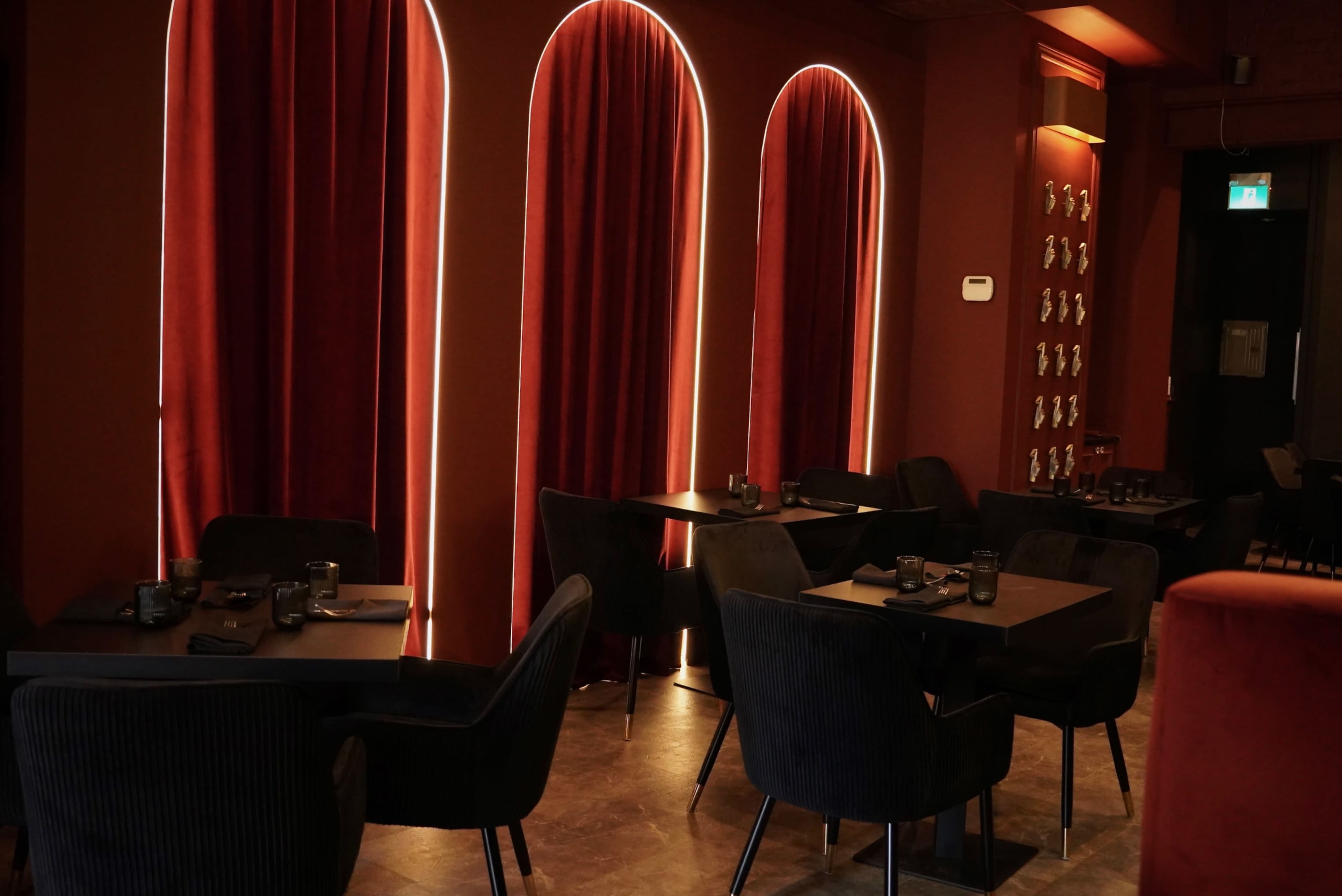 The interior of a restaurant features dark velvet chairs and tables arranged beneath large, arched windows framed with red curtains.