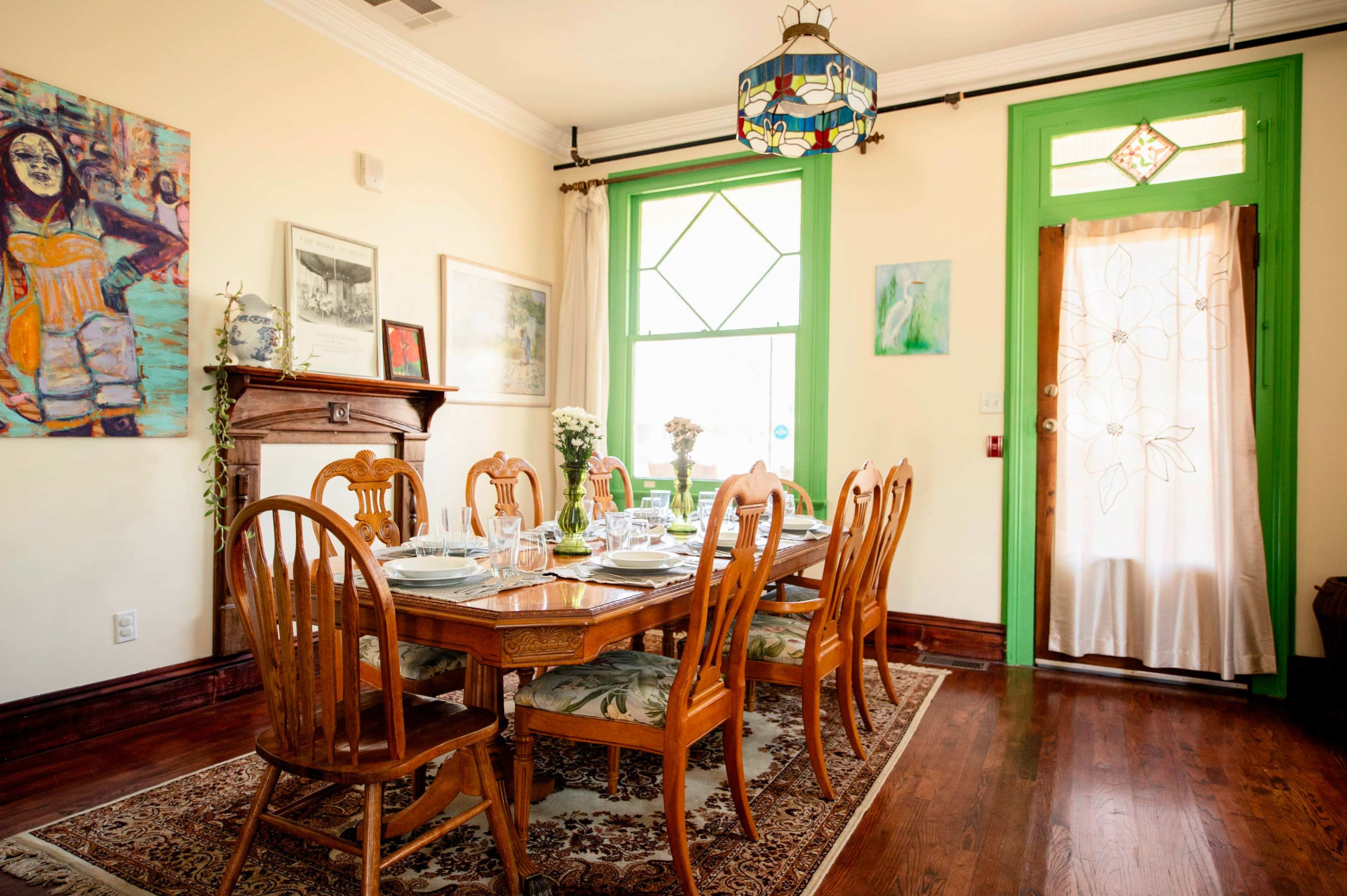 The image shows a dining room with a large wooden table set for a meal, surrounded by wooden chairs, and featuring colorful artwork and a bright green door.