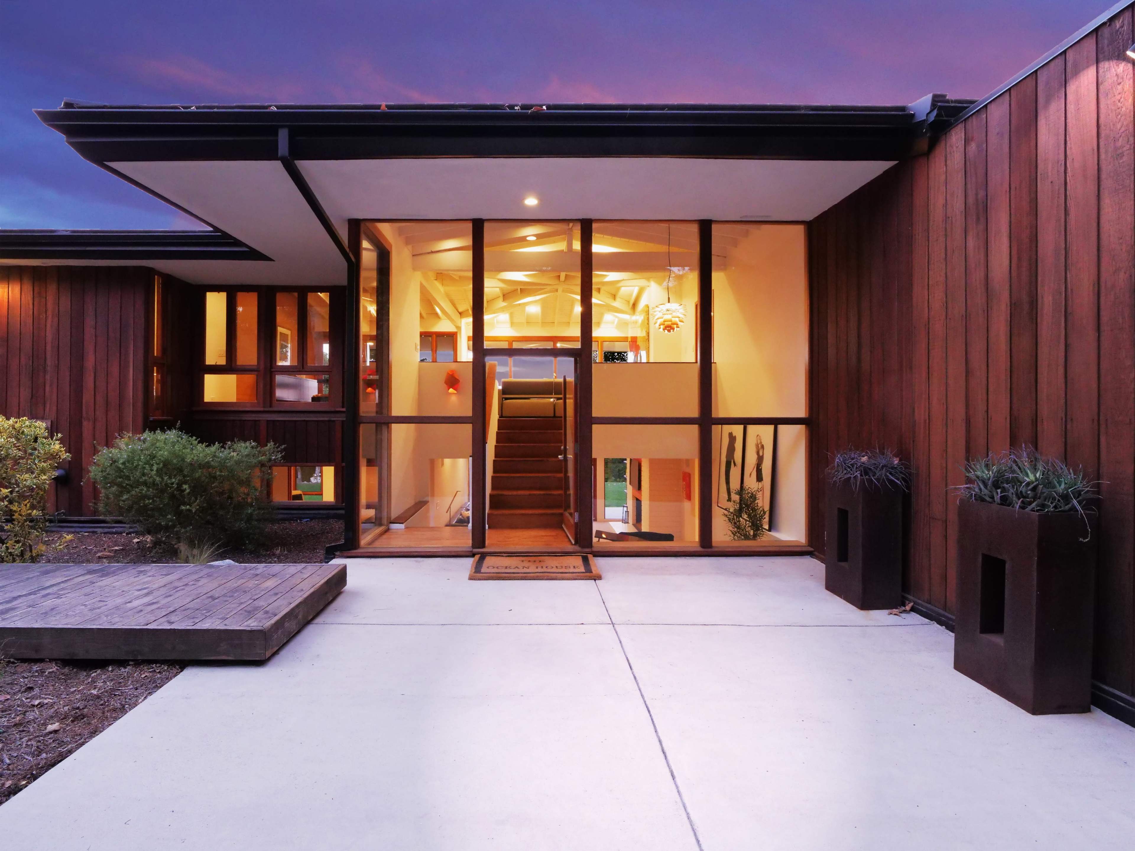 The image shows a modern house entrance with a wooden façade and large glass panels, leading to an interior space illuminated by warm lighting.