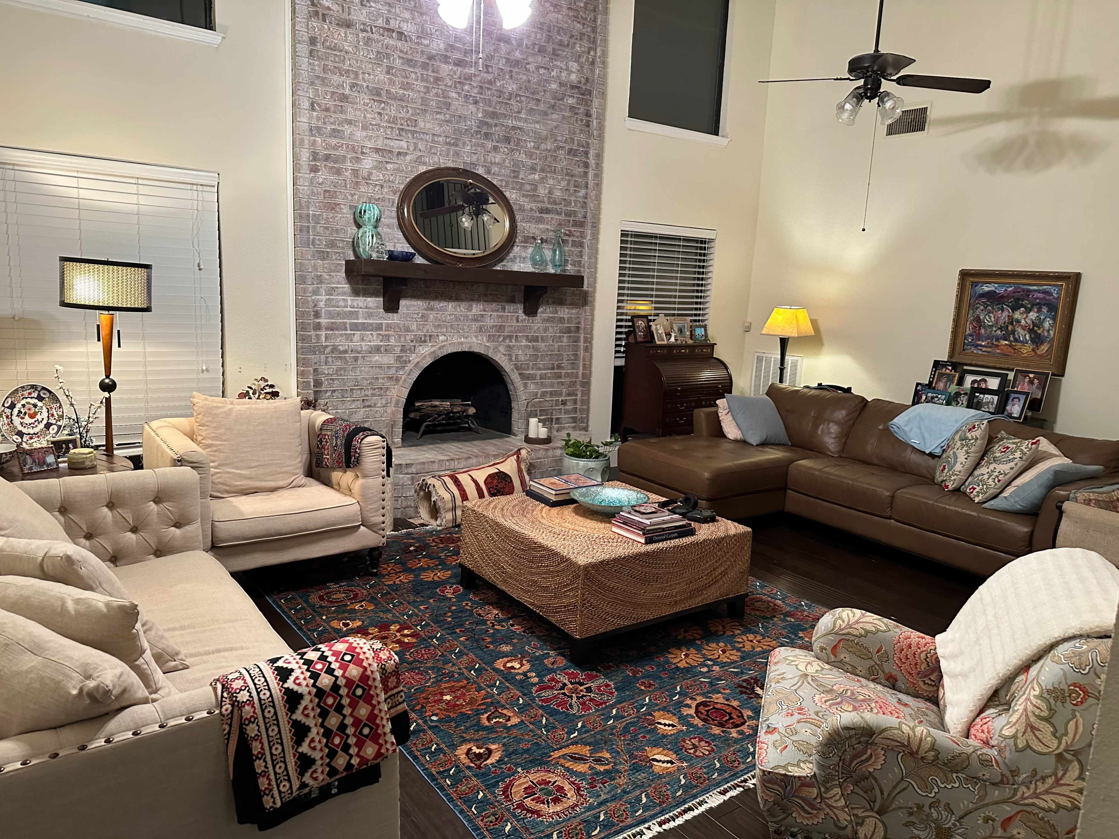 A living room features a large brick fireplace with a round mirror above it, surrounded by two sofas, an armchair, and a coffee table on a patterned rug.