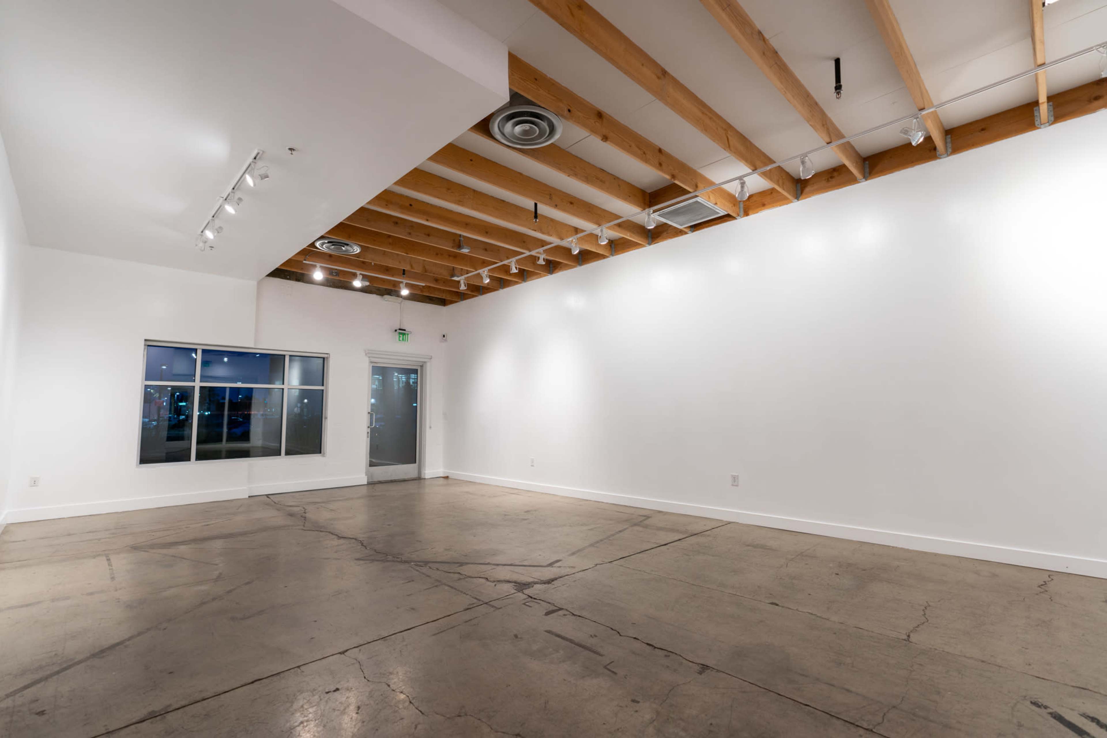 A spacious, empty room with a polished concrete floor and exposed wooden beams in the ceiling, featuring large windows and a glass door.