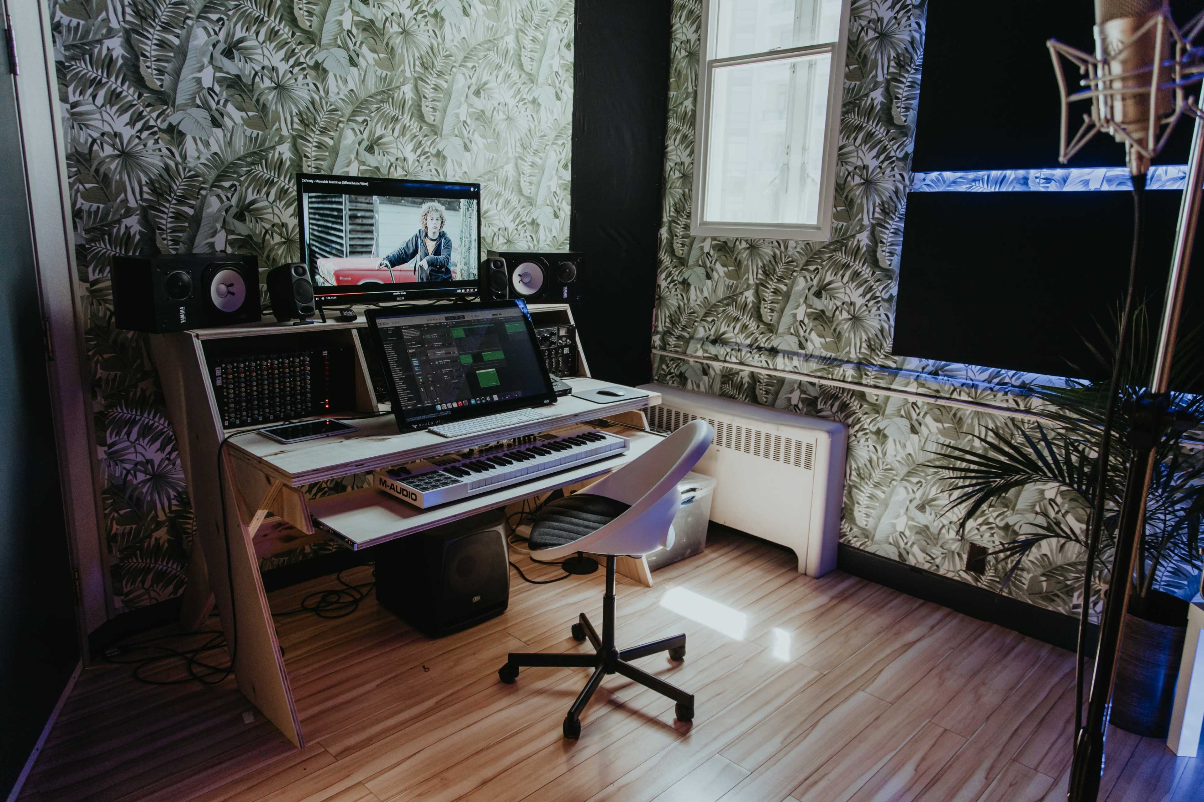 A modern music studio features a keyboard, computer monitor, and audio equipment against a leafy wallpaper backdrop.