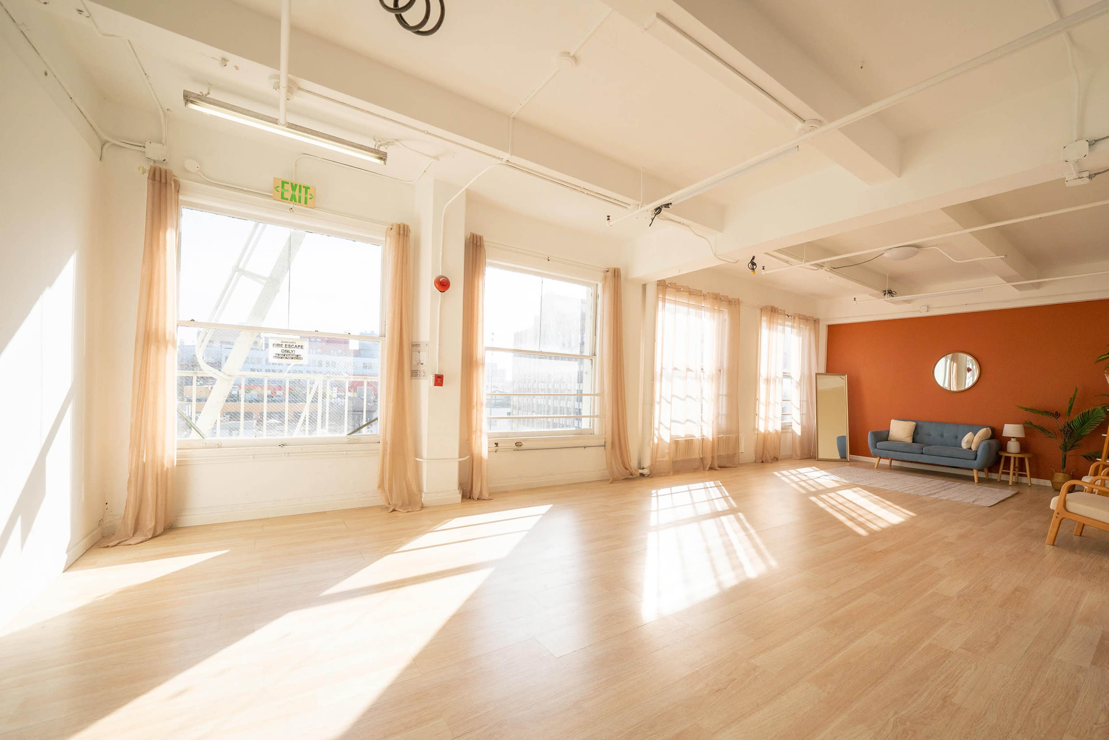 The image shows a bright, empty room with large windows, light wooden flooring, and a seating area featuring a blue couch and an orange accent wall.