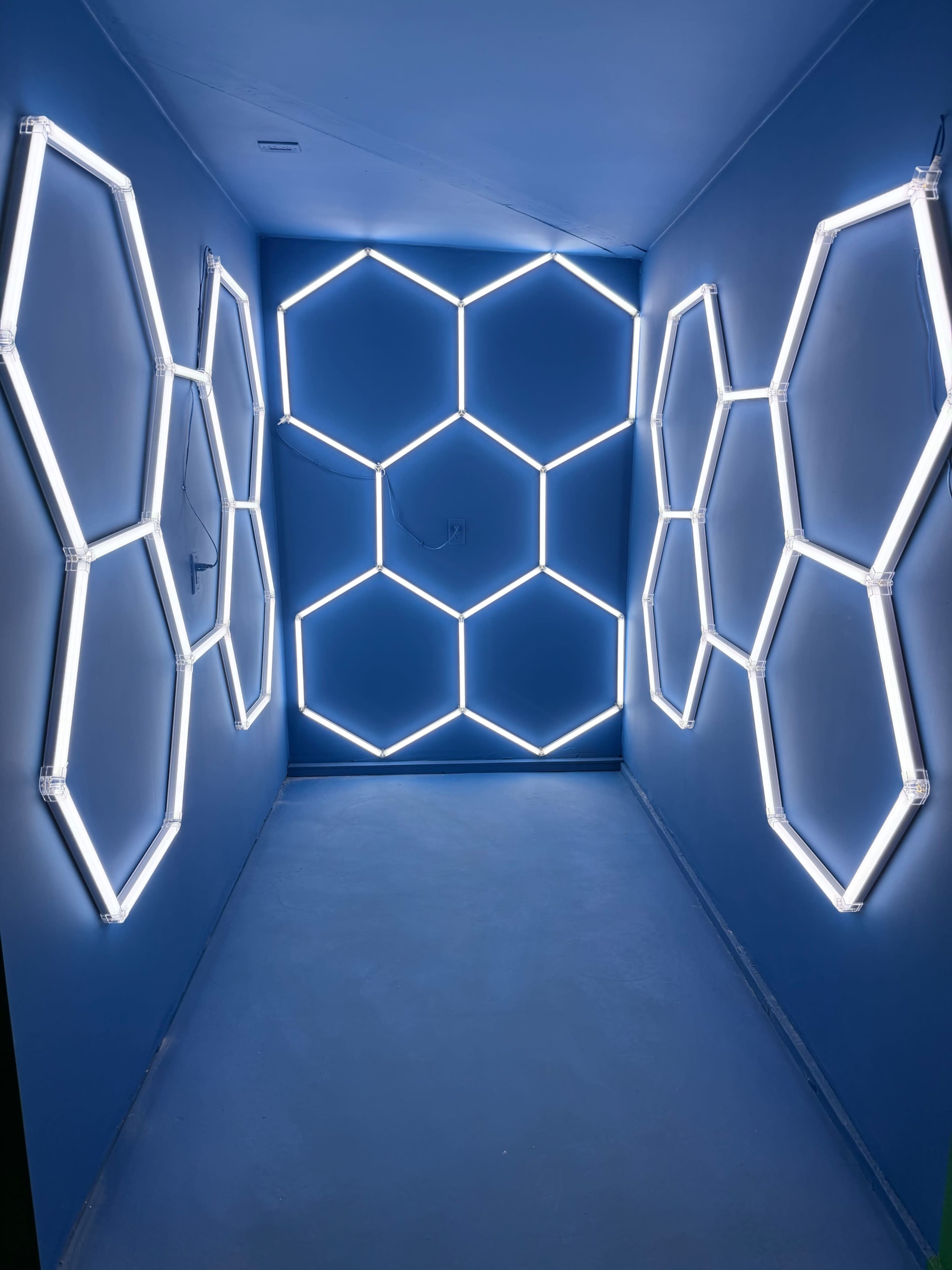 The image shows a narrow hallway with blue walls and illuminated hexagonal neon light fixtures arranged on both sides.