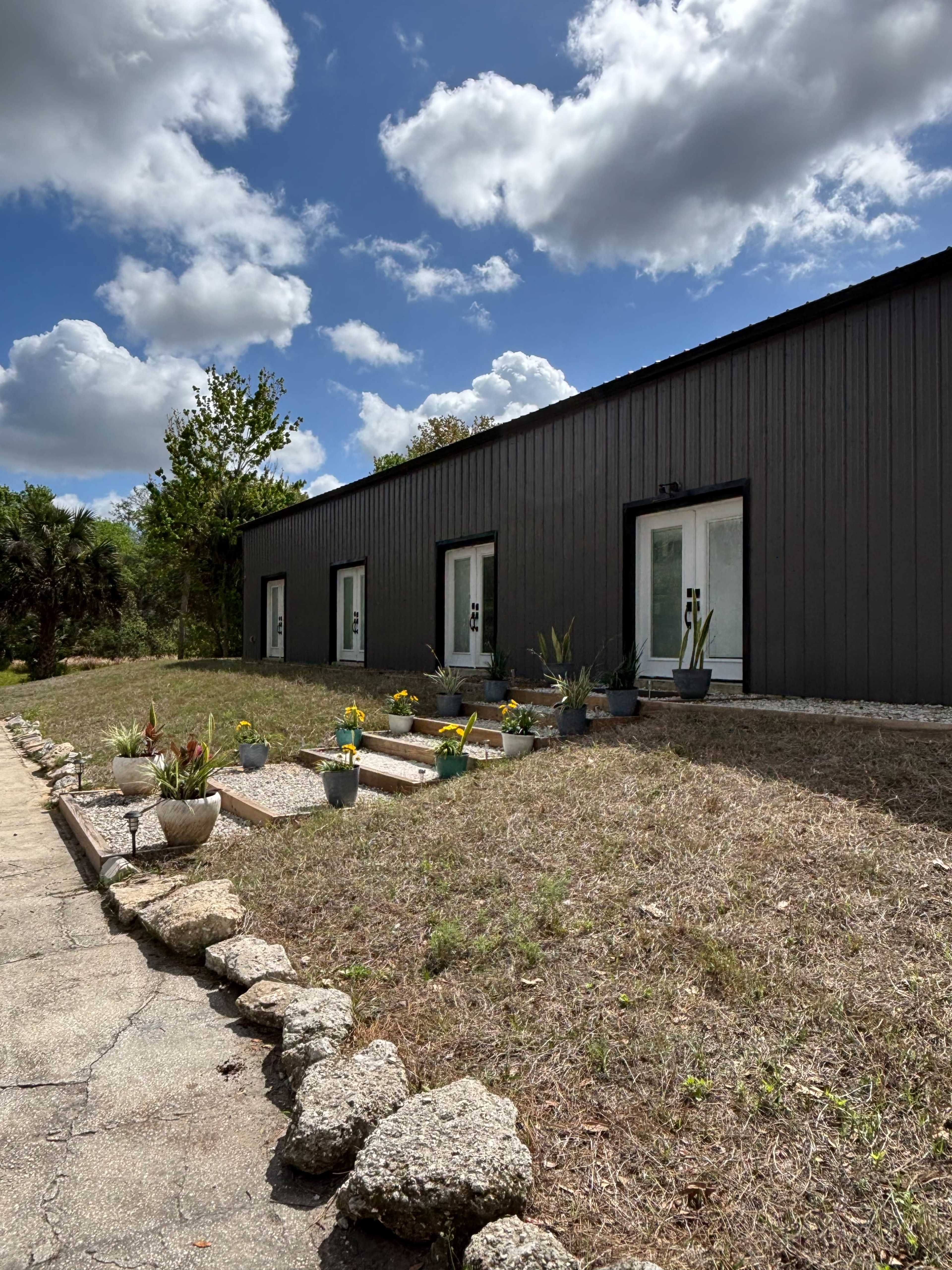 A modern building with a dark exterior features three large windows, a pathway lined with rocks, and flower pots in a grassy area under a partly cloudy sky.
