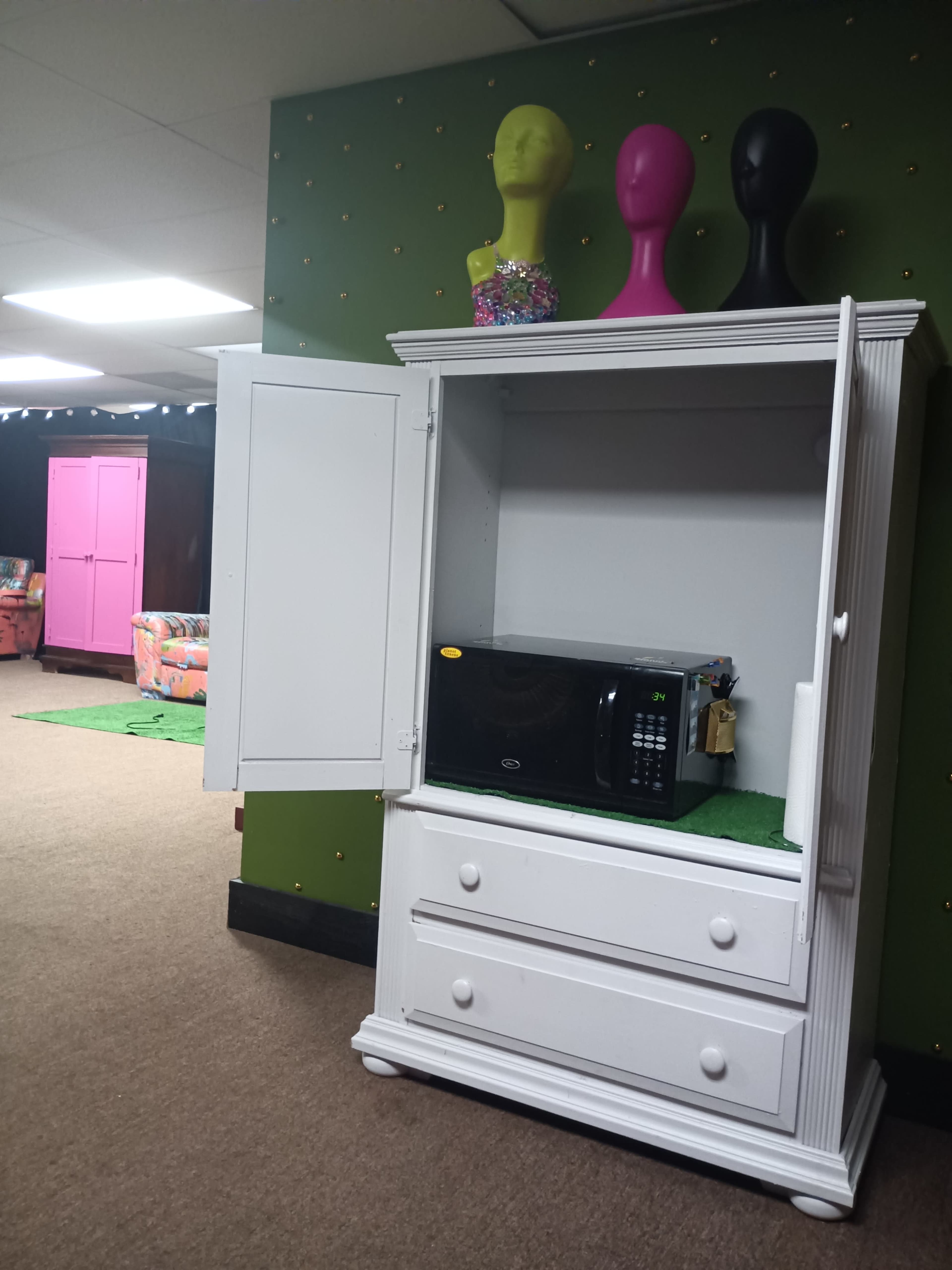The image shows a white armoire with its doors open, revealing a microwave inside, set against a green wall decorated with colorful accents.