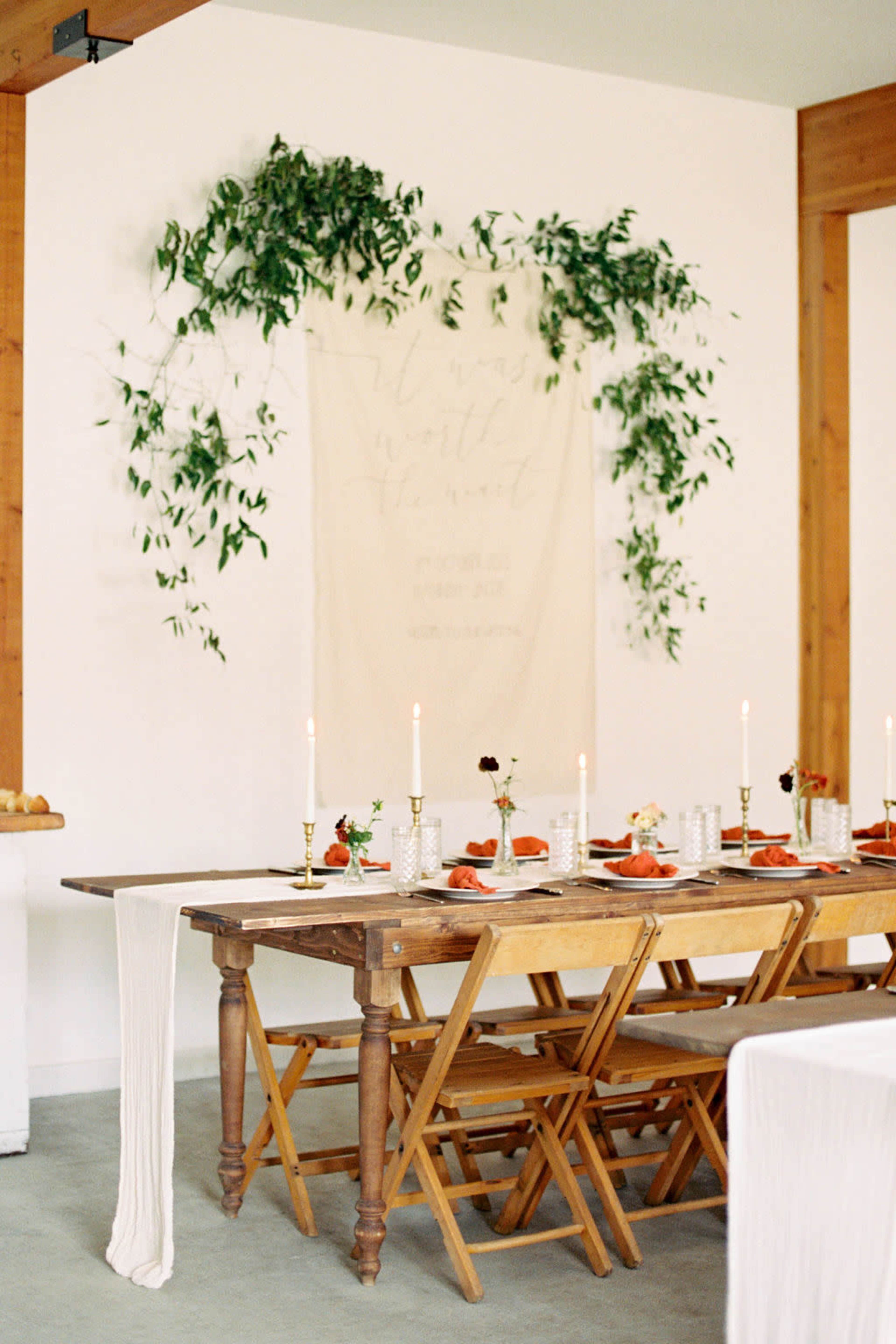 A long wooden dining table is elegantly set with white tableware and candles against a backdrop of a wall adorned with green foliage and a decorative fabric hanging.
