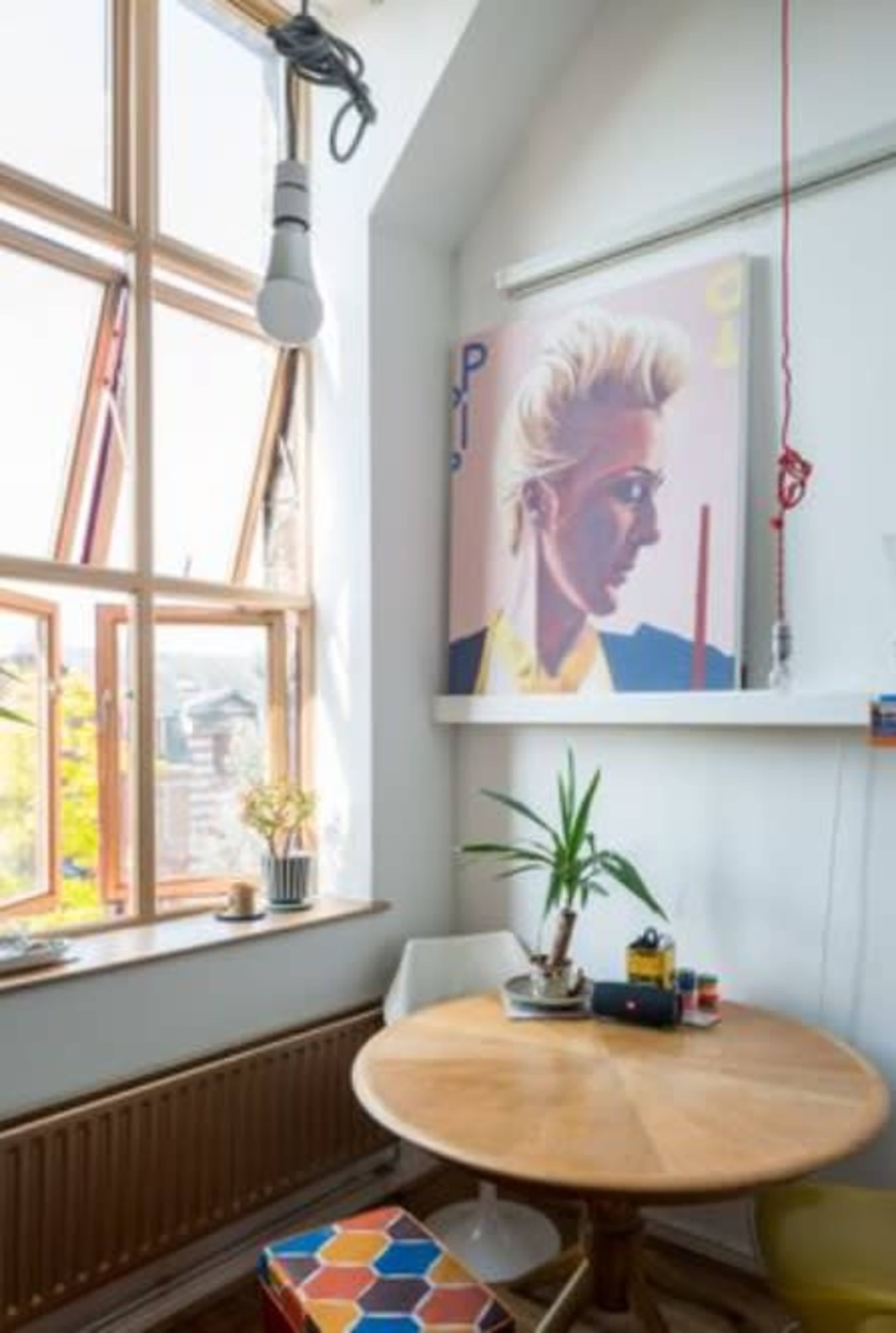 A corner of a room features a round wooden table, a potted plant, and a large poster depicting a profile of a woman, with large windows allowing natural light to enter.