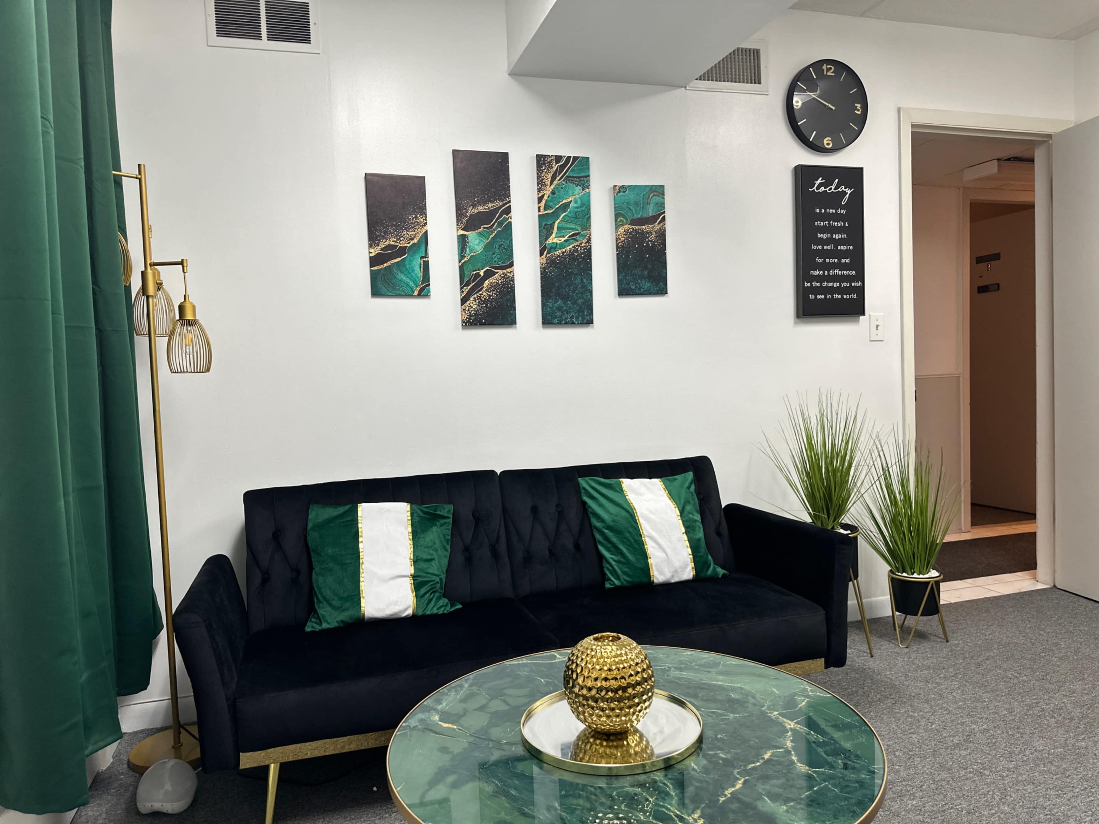 A modern living room with a black couch adorned with green and white pillows, a round glass coffee table, wall art featuring abstract designs, and a wall clock.