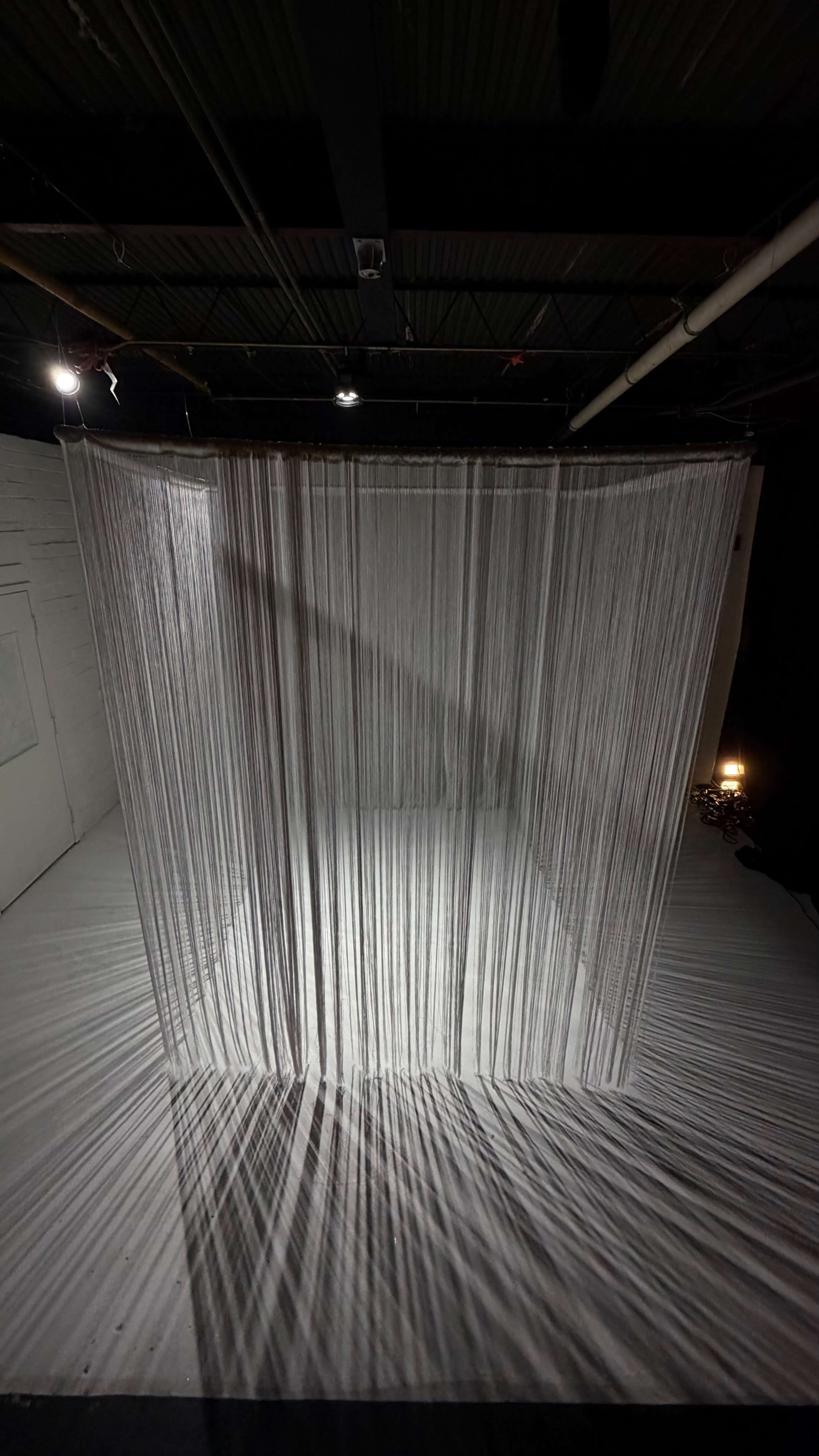The image shows a white curtain made of thin strings draping in a semi-circular formation in a dimly lit room.