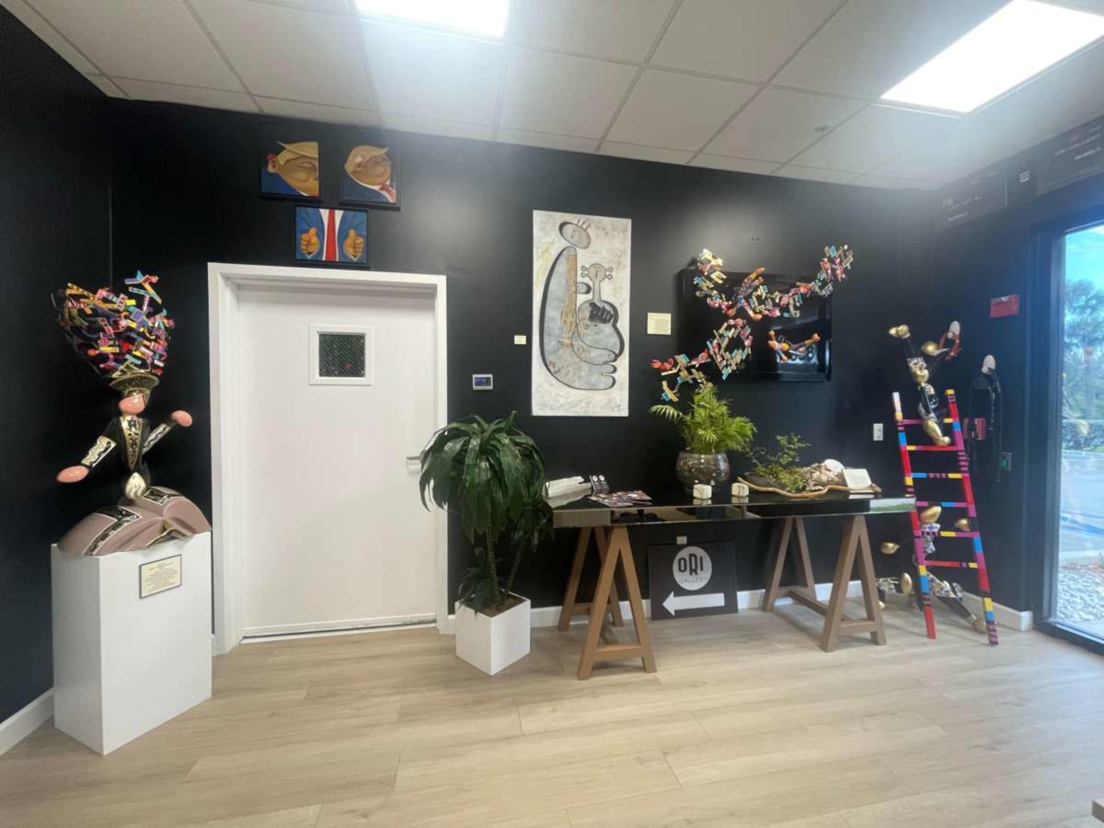The image depicts an art-filled interior space featuring colorful sculptures and artwork on black walls, with a wooden table in the center displaying decorative items and a green plant.