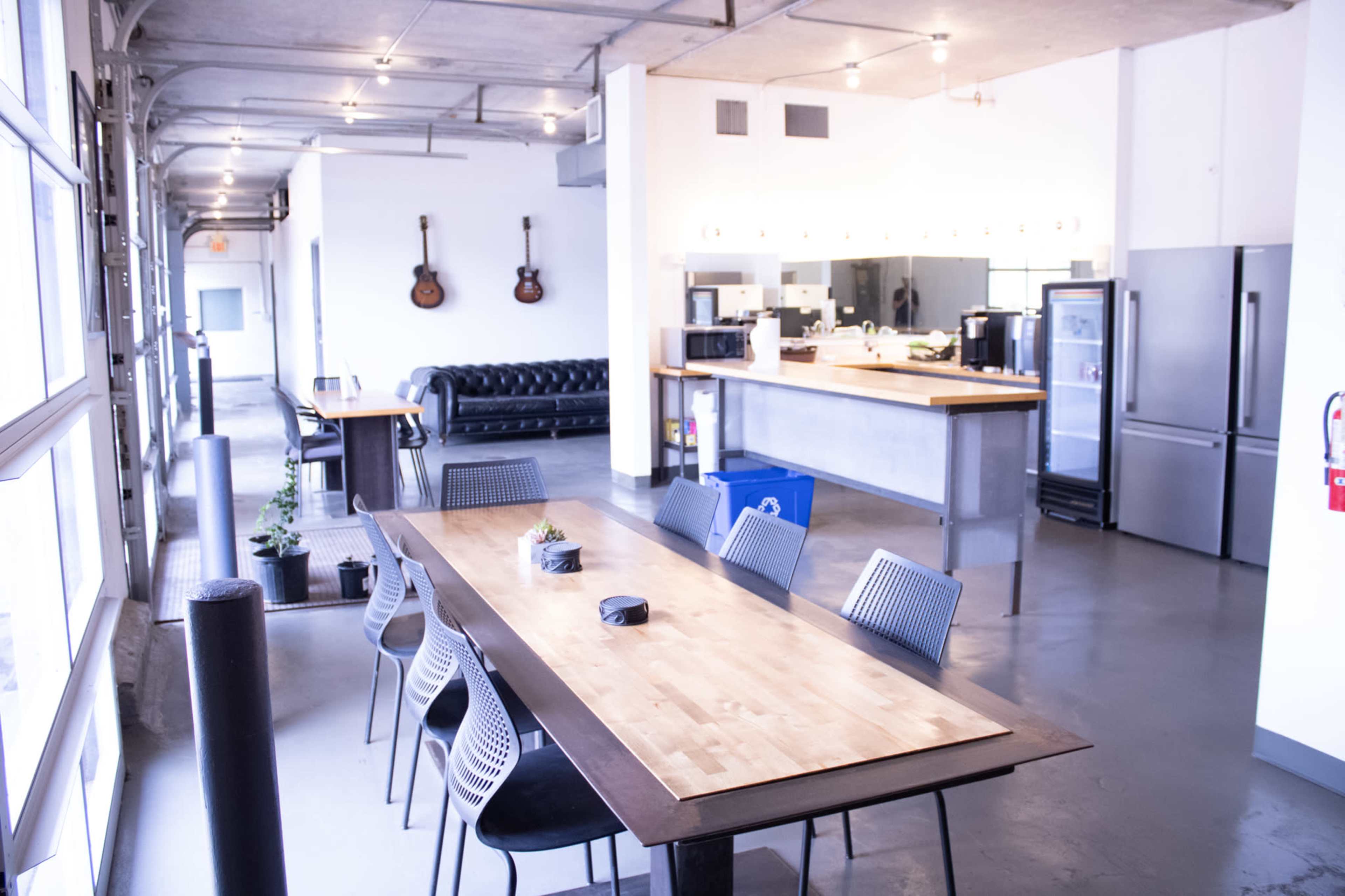 The image shows a modern, open-concept communal space with a long wooden table, black chairs, a kitchen area, and a seating area featuring a black couch and acoustic guitars on the wall.
