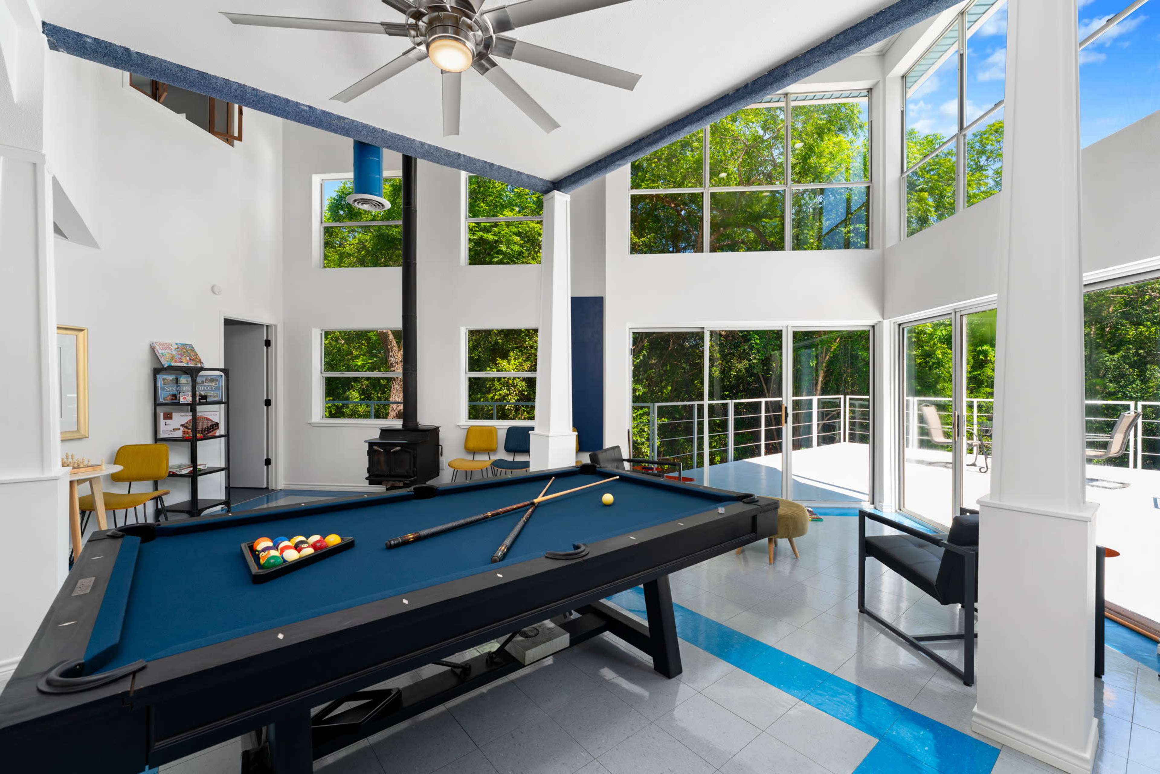 The image shows a modern game room featuring a billiards table, large windows, and a wood stove.