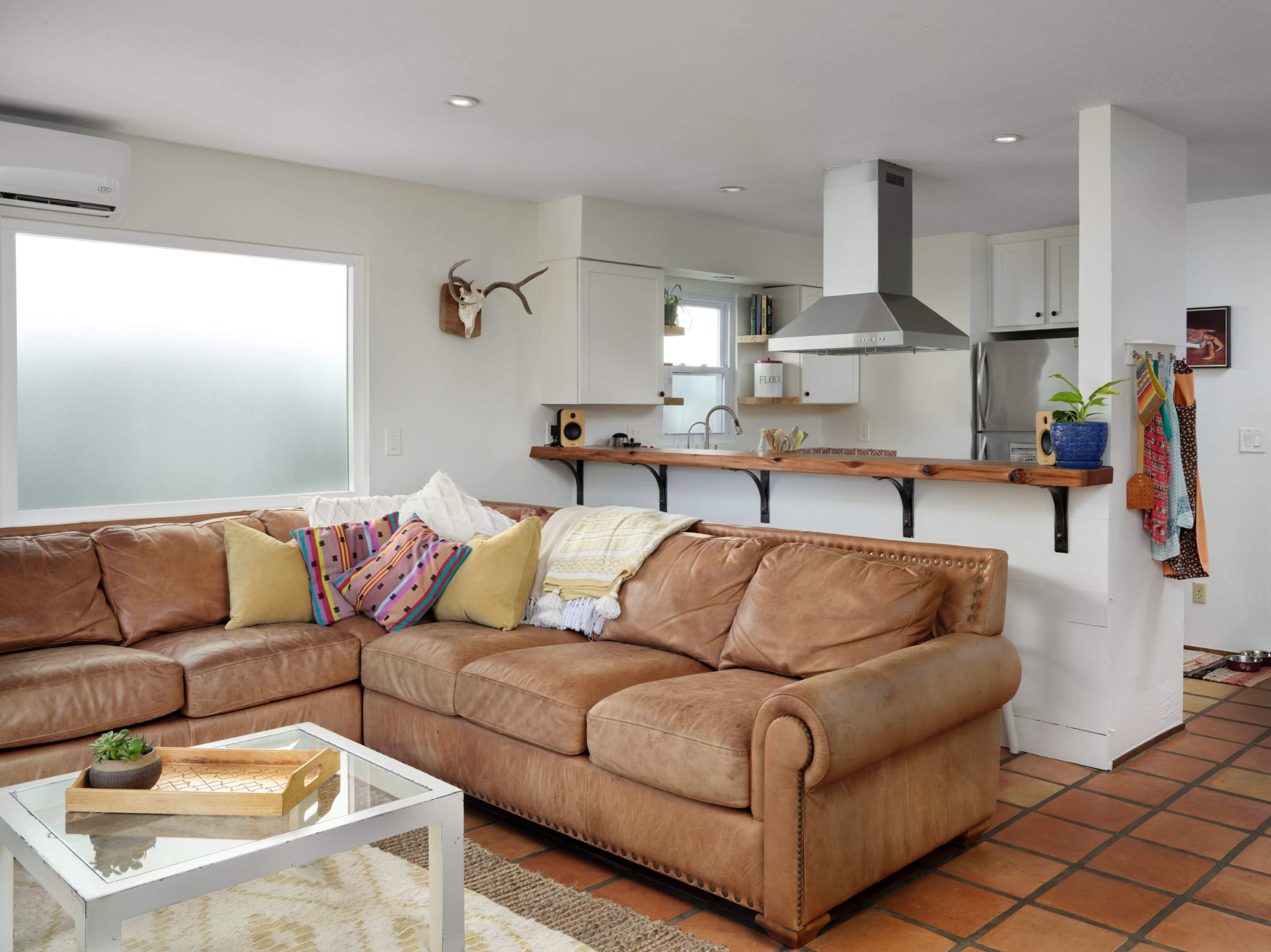 A spacious living room features a large brown leather sectional sofa, a glass coffee table, and an open kitchen area with modern appliances.