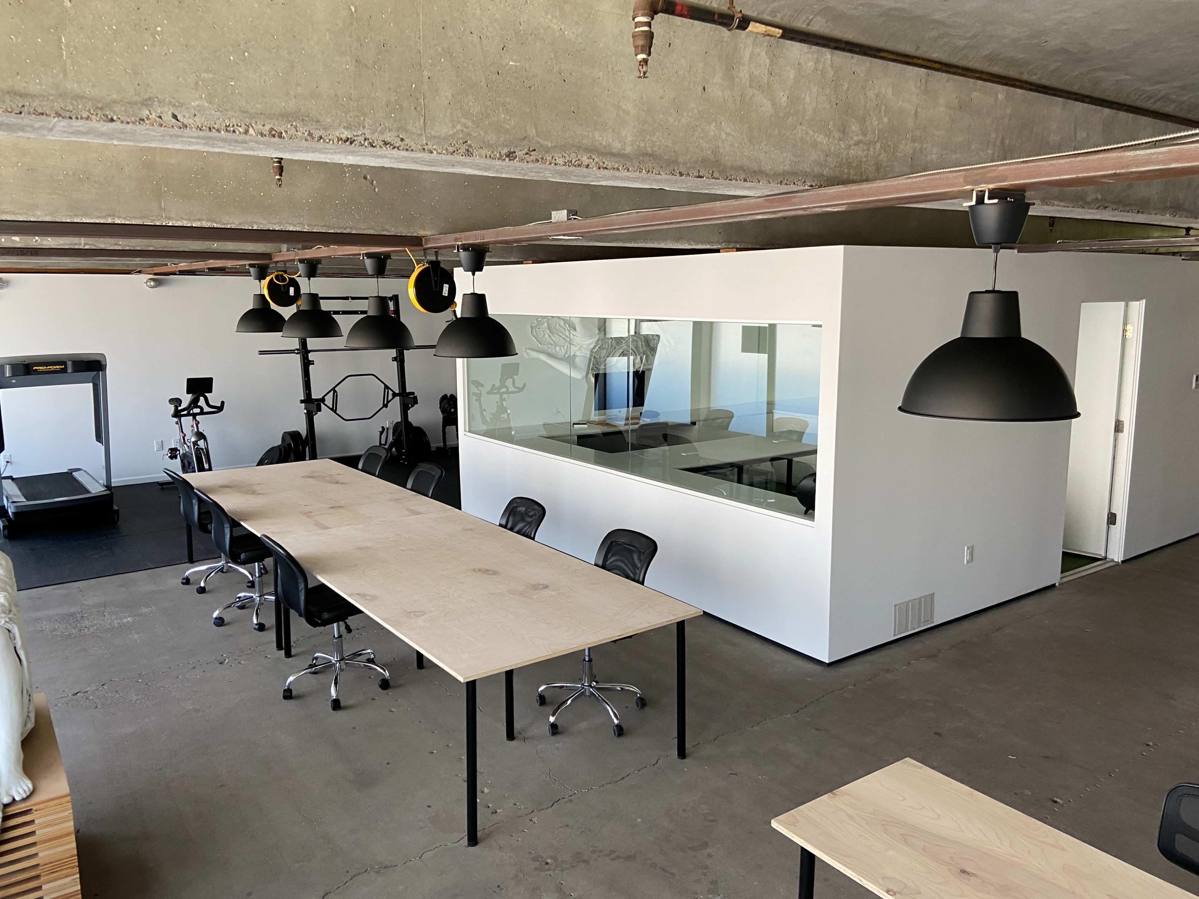 A modern office space featuring a large table with chairs, a treadmill in the background, and a glass-walled meeting room.