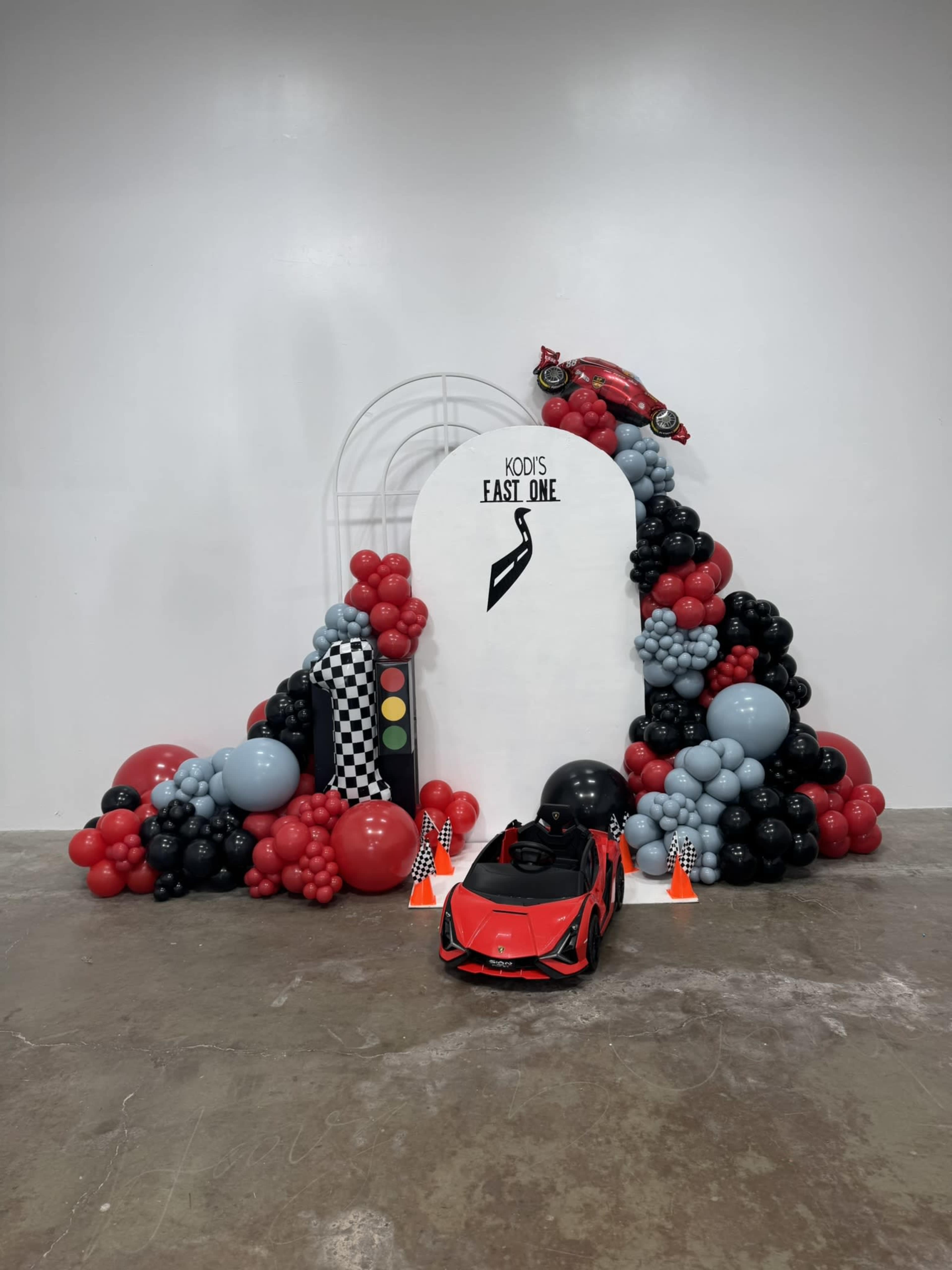 The scene features a decorative balloon display in red, black, and gray, with a small toy race car parked in front of a white arch that reads "KOD'S EAST ONE."