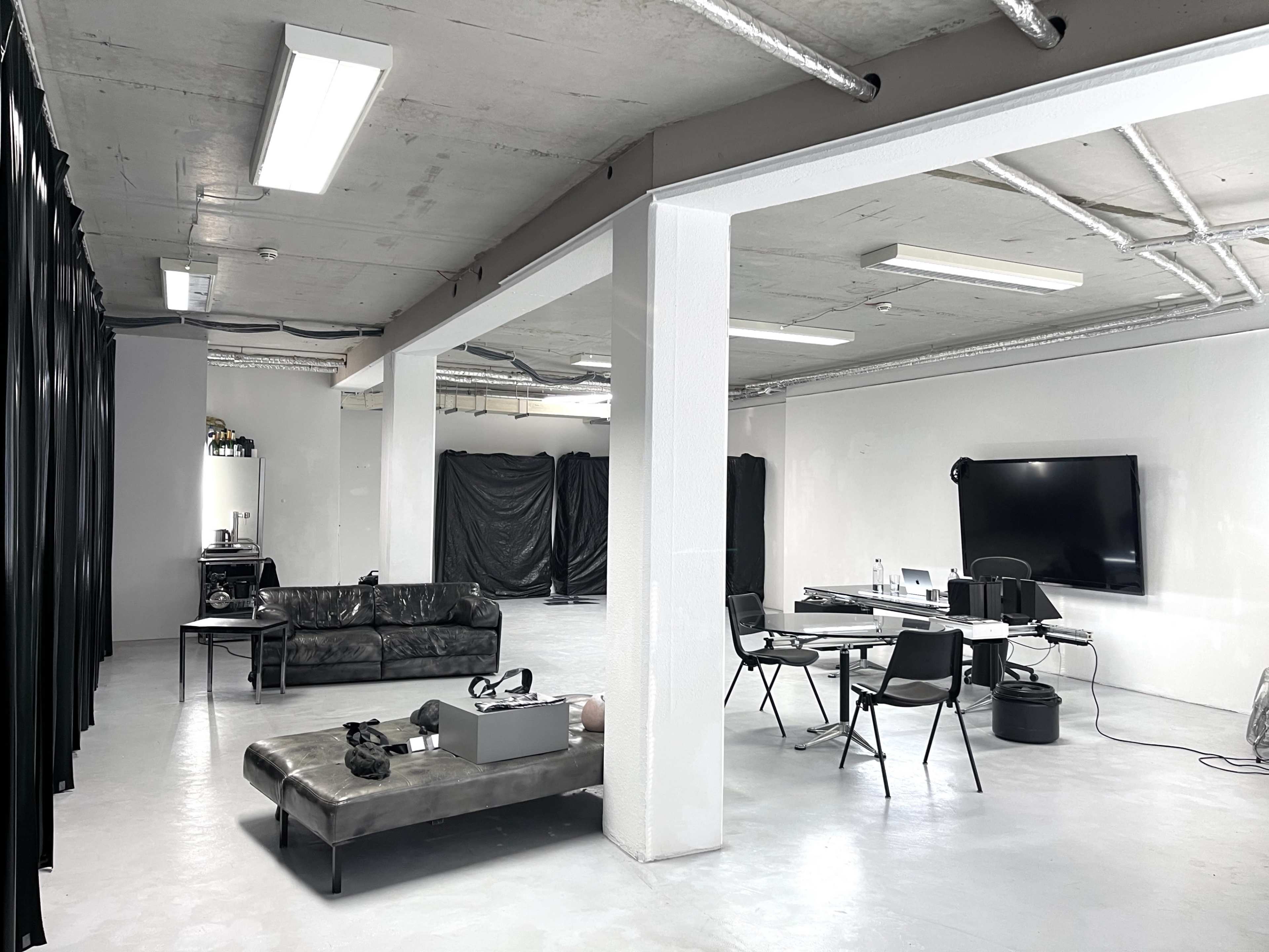 The image shows a spacious, modern studio with a black couch, a coffee table, a set of chairs around a desk, and a large screen on one wall.