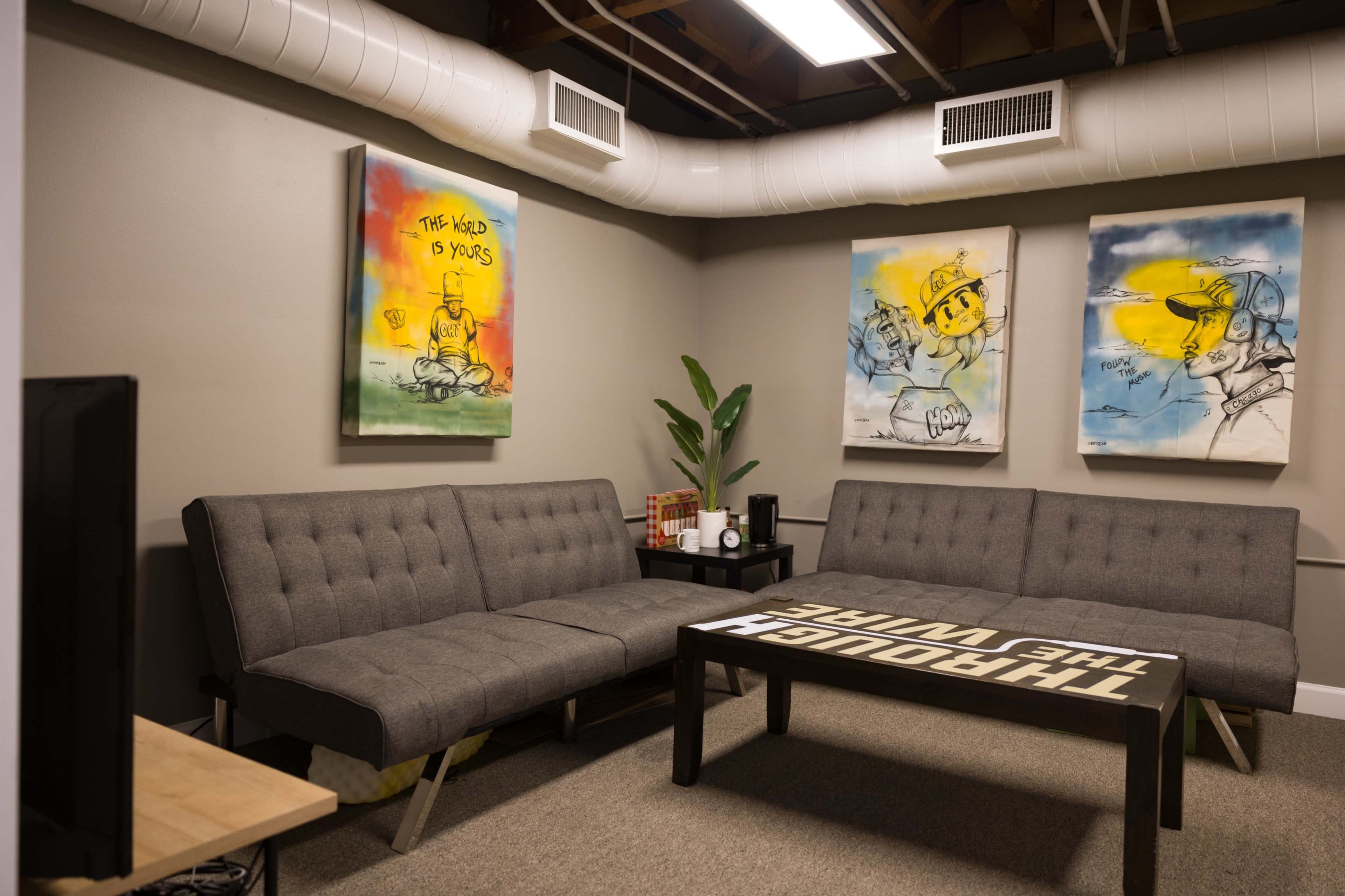 A casual lounge area with a gray sectional sofa, a coffee table printed with the phrase "THROUGH THE WIRE," and three colorful art pieces on the walls.