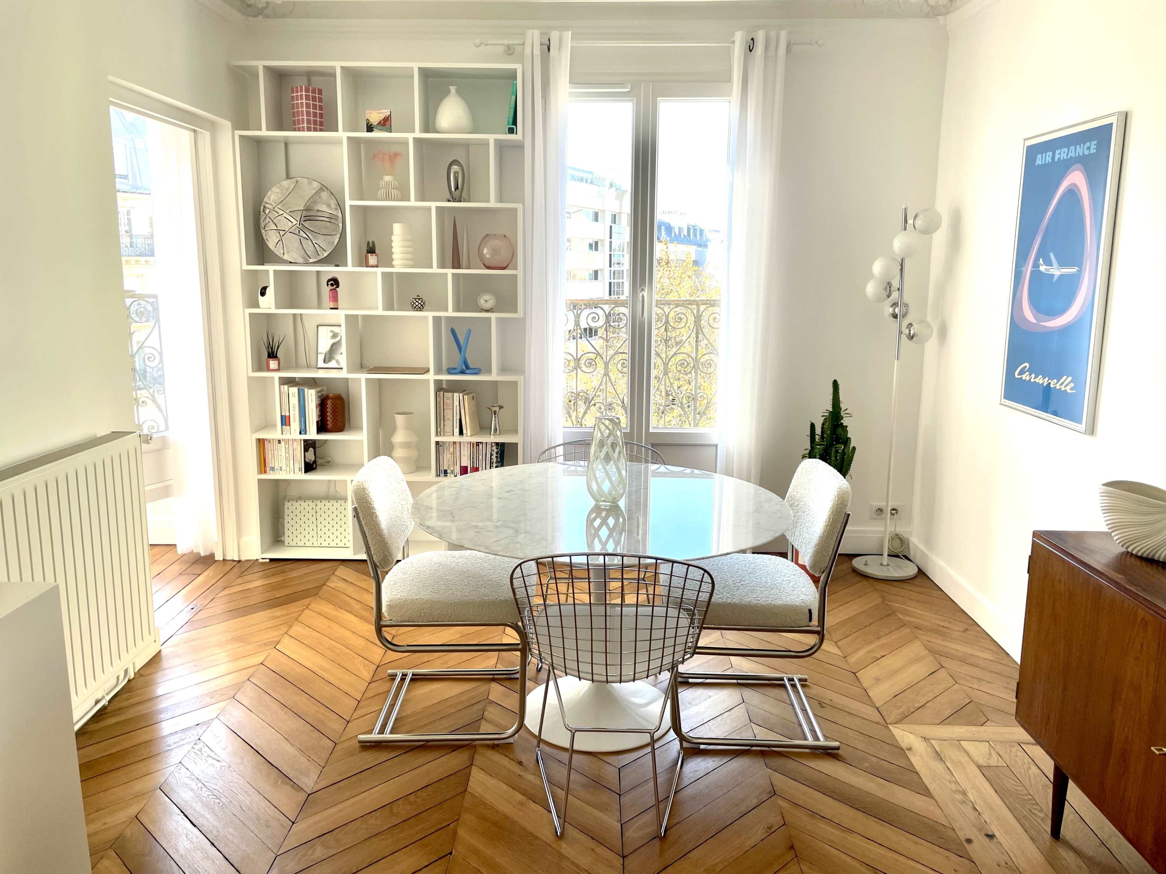 A modern dining area features a round table surrounded by wire chairs, hardwood flooring, and a bookshelf filled with decor and books.