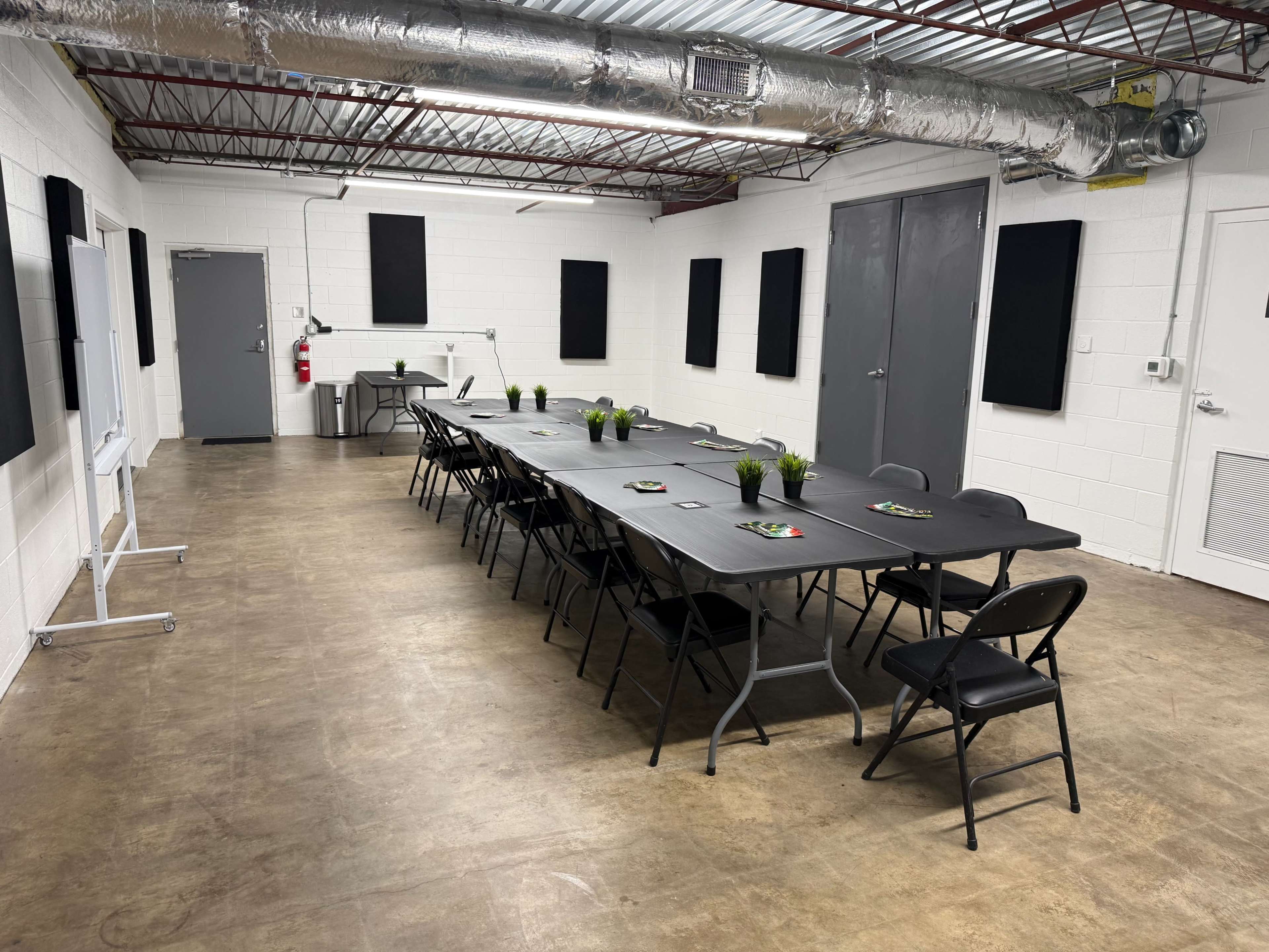 The image shows a spacious meeting room with a long table set with small plants and placemats, surrounded by chairs, and featuring white walls and a concrete floor.