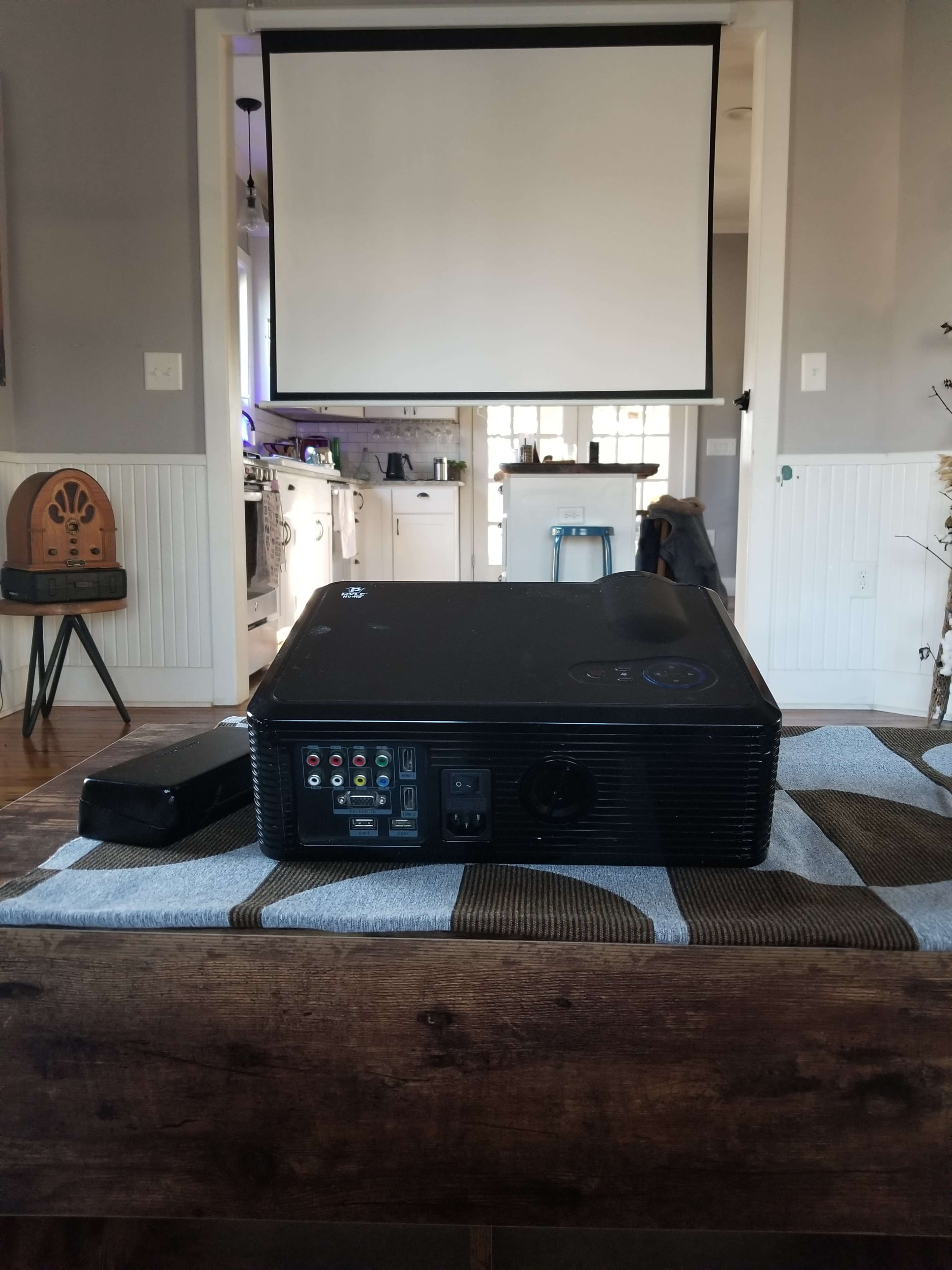 A projector is placed on a wooden table in a room with a large screen in the background.