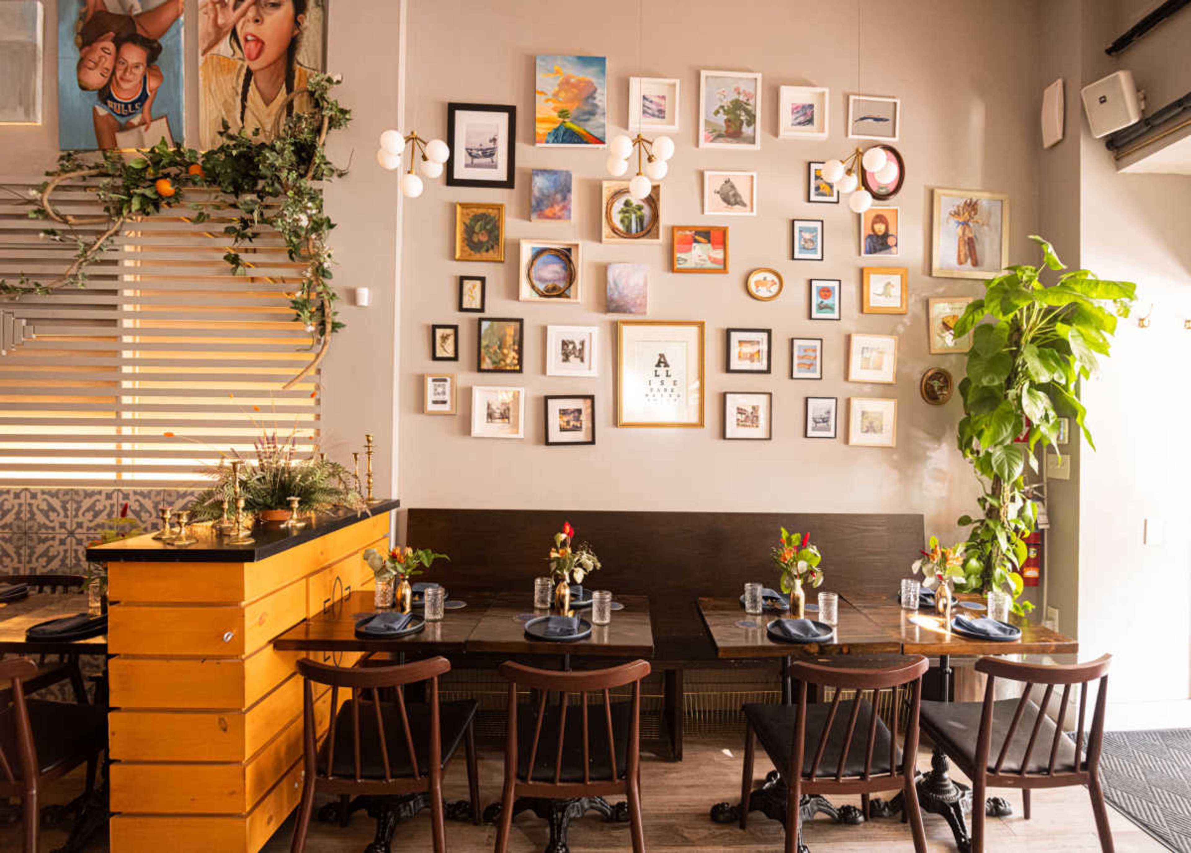 The image shows a dining area with wooden tables and chairs, decorated with a gallery wall featuring various framed pictures and a plant beside the tables.