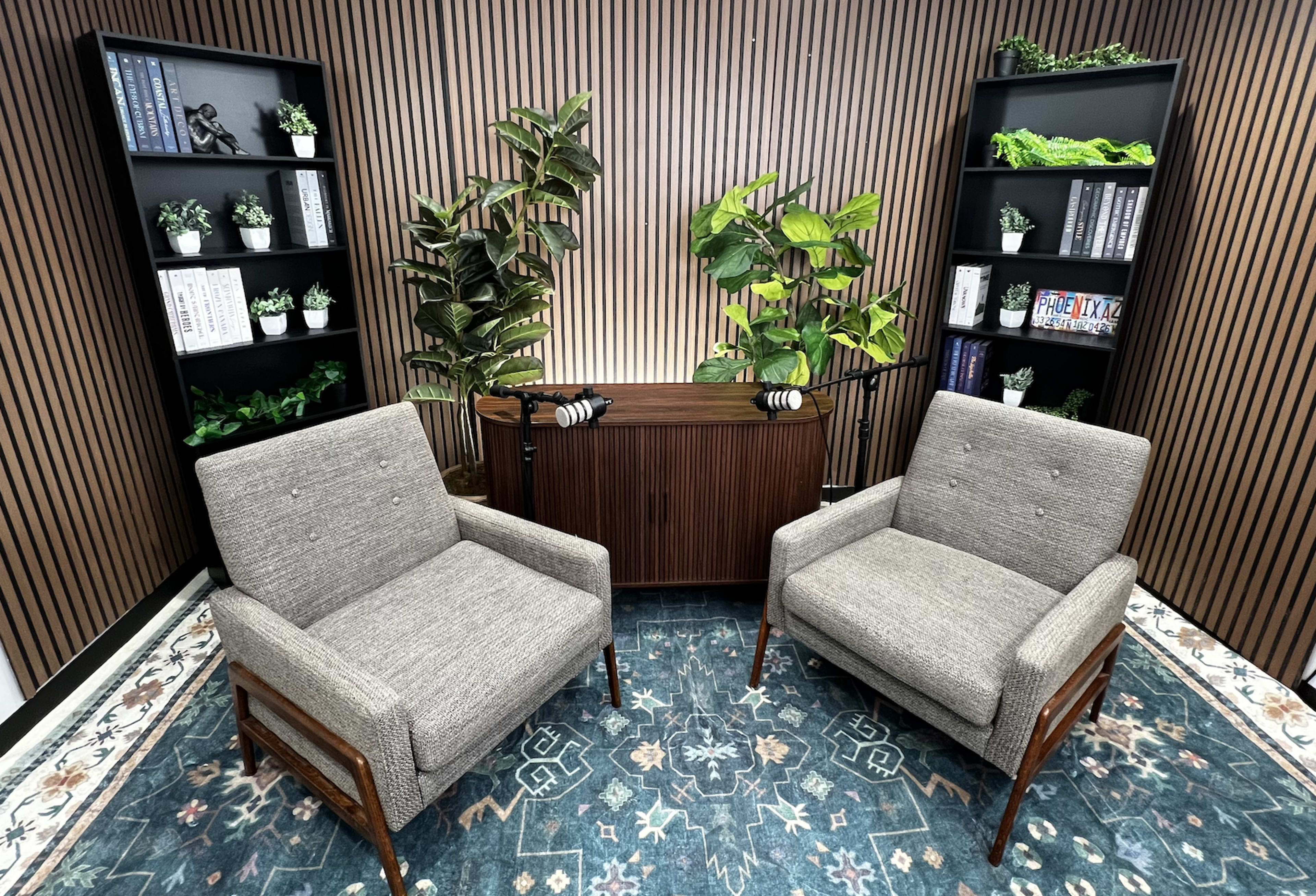 The image shows a cozy discussion space featuring two gray armchairs facing a wooden table, with shelves of plants and books on either side against a striped wooden wall.