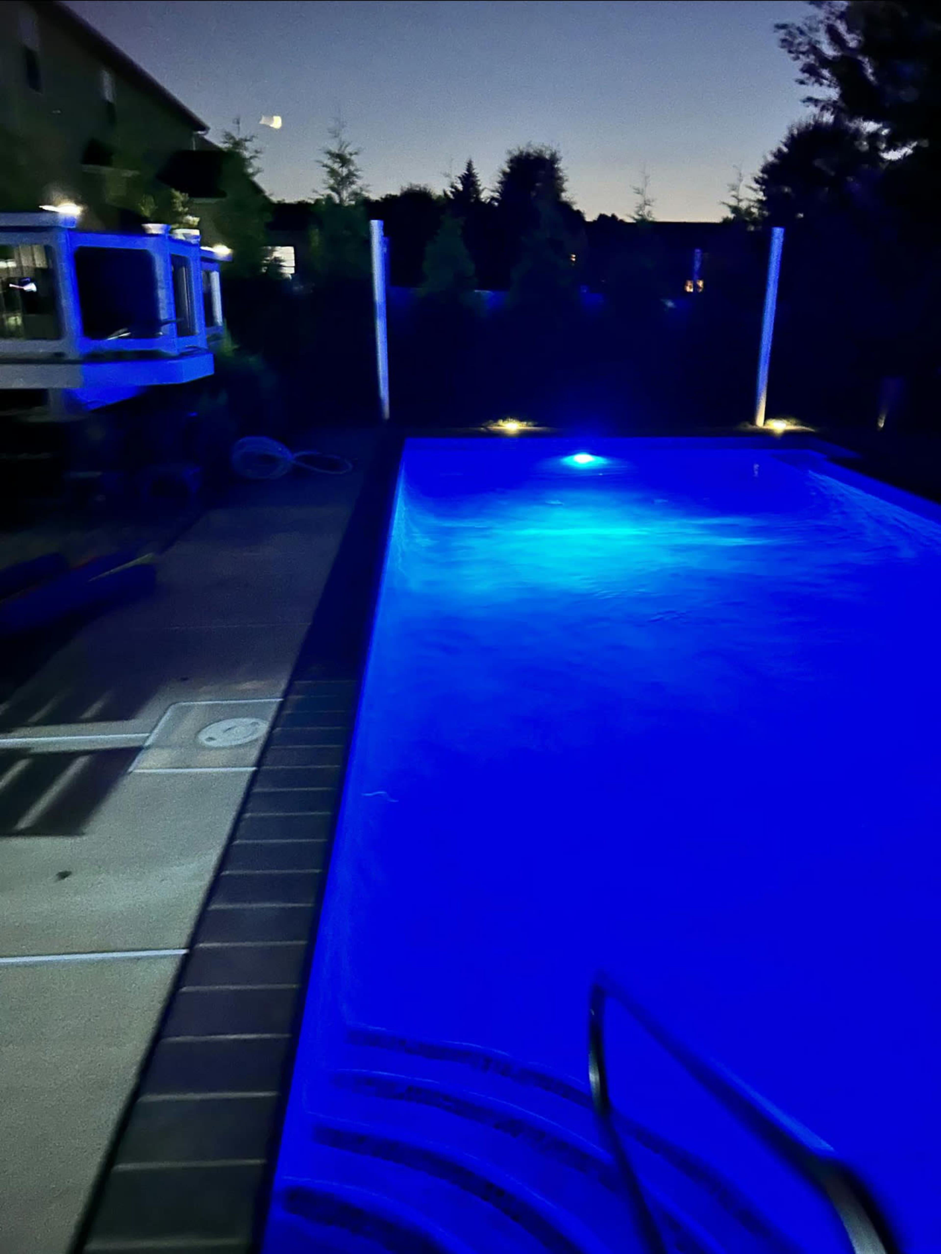 A swimming pool with blue lighting is surrounded by trees and a building at dusk.