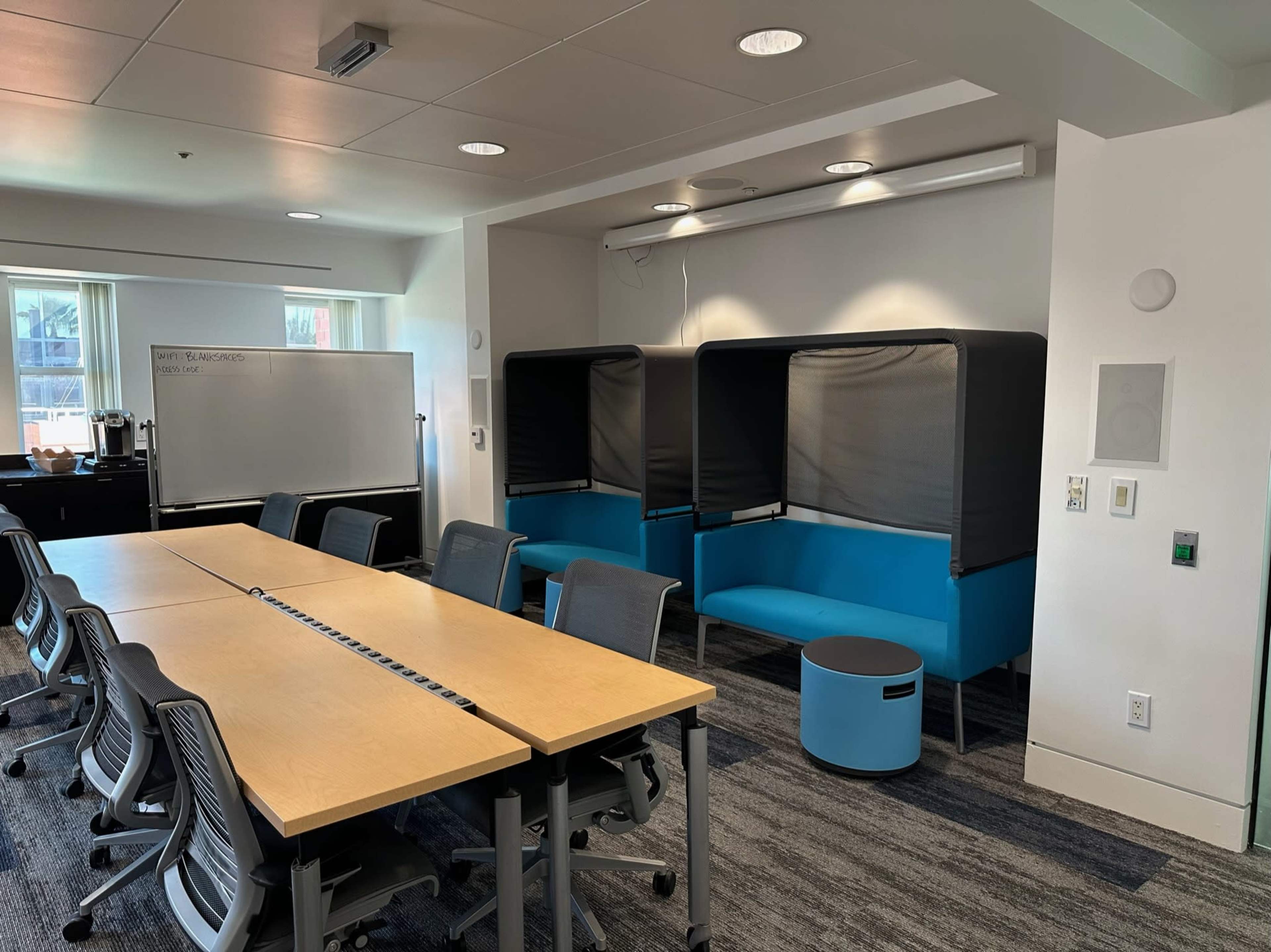 A modern office space with a long table, ergonomic chairs, and two seating pods along the wall.
