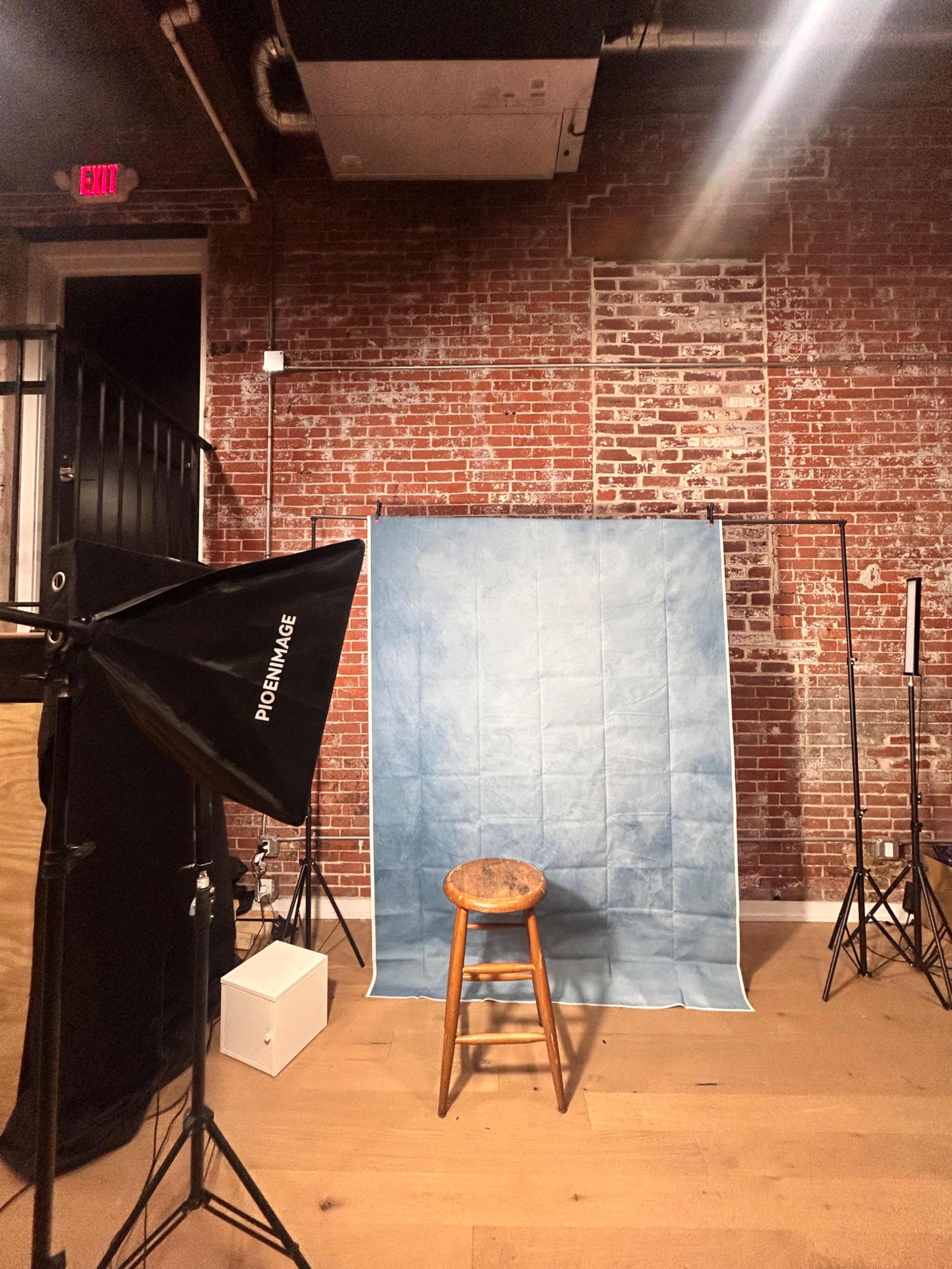 The image shows a photography studio with a blue backdrop, a wooden stool in front of it, and studio lighting equipment arranged around the space.