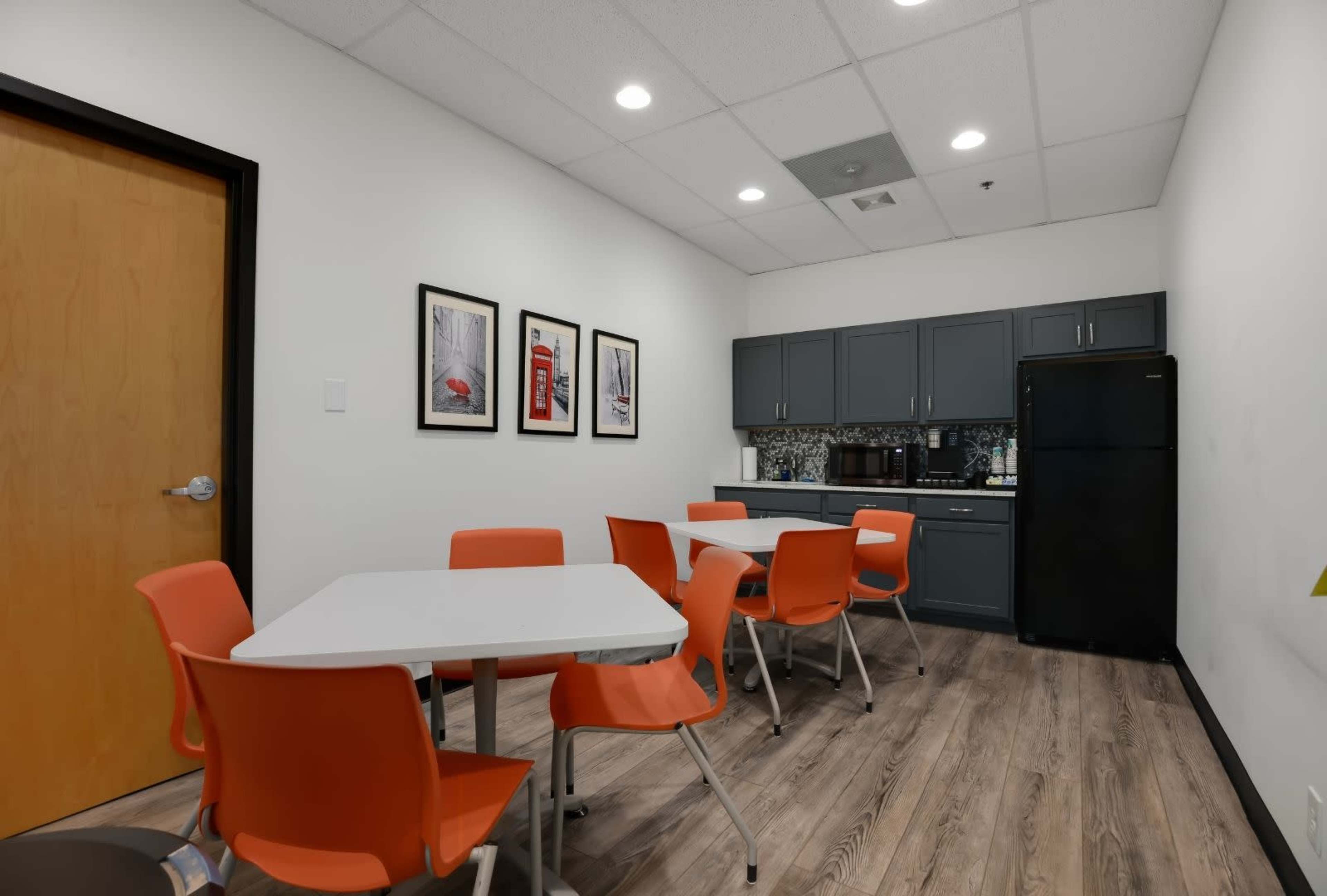 A break room features a table surrounded by orange chairs, with a dark cabinet and refrigerator, and framed art on the wall.