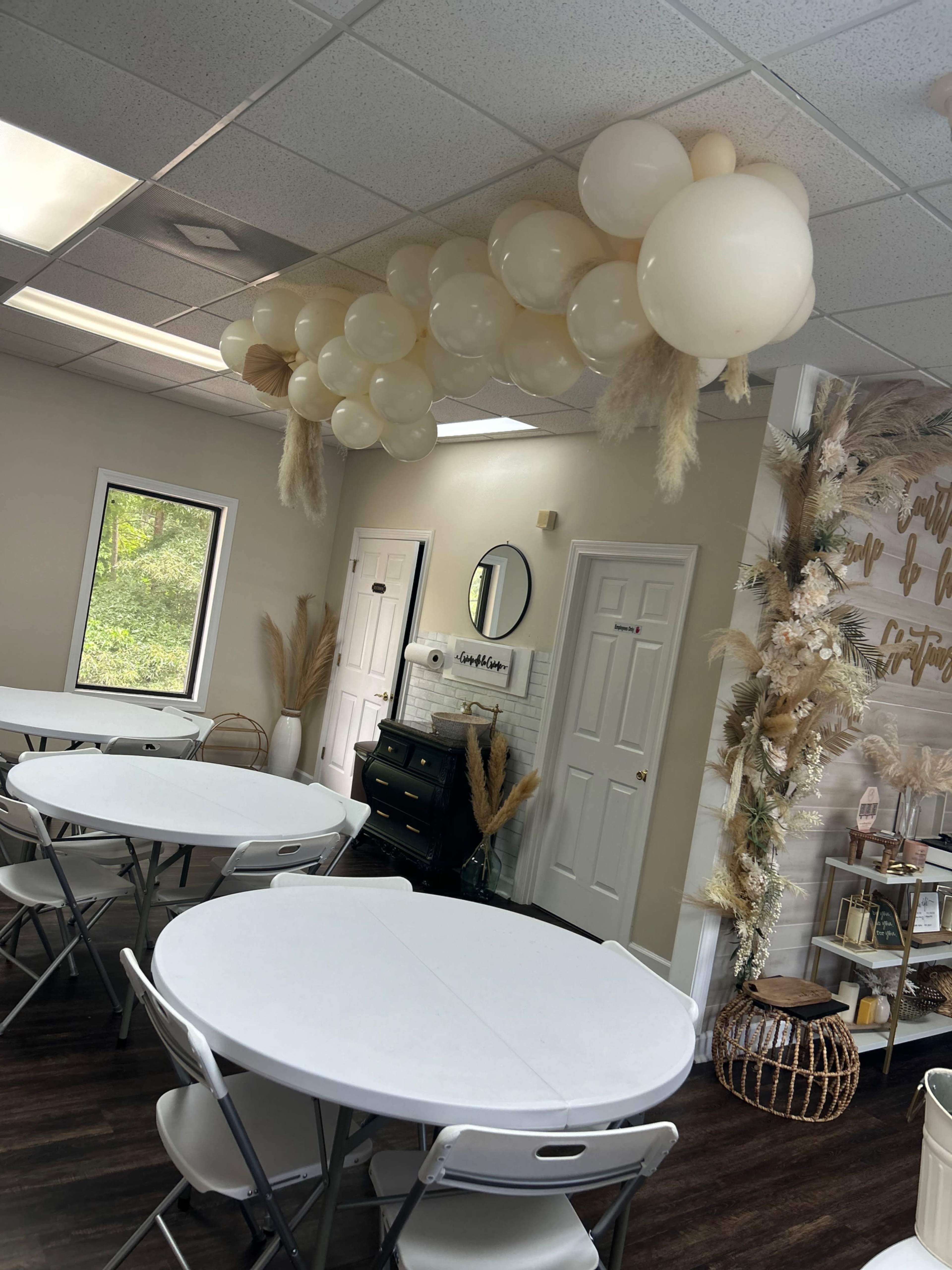 The image shows a decorated indoor event space with white tables and a balloon arrangement overhead, complemented by dried floral accents on the wall.