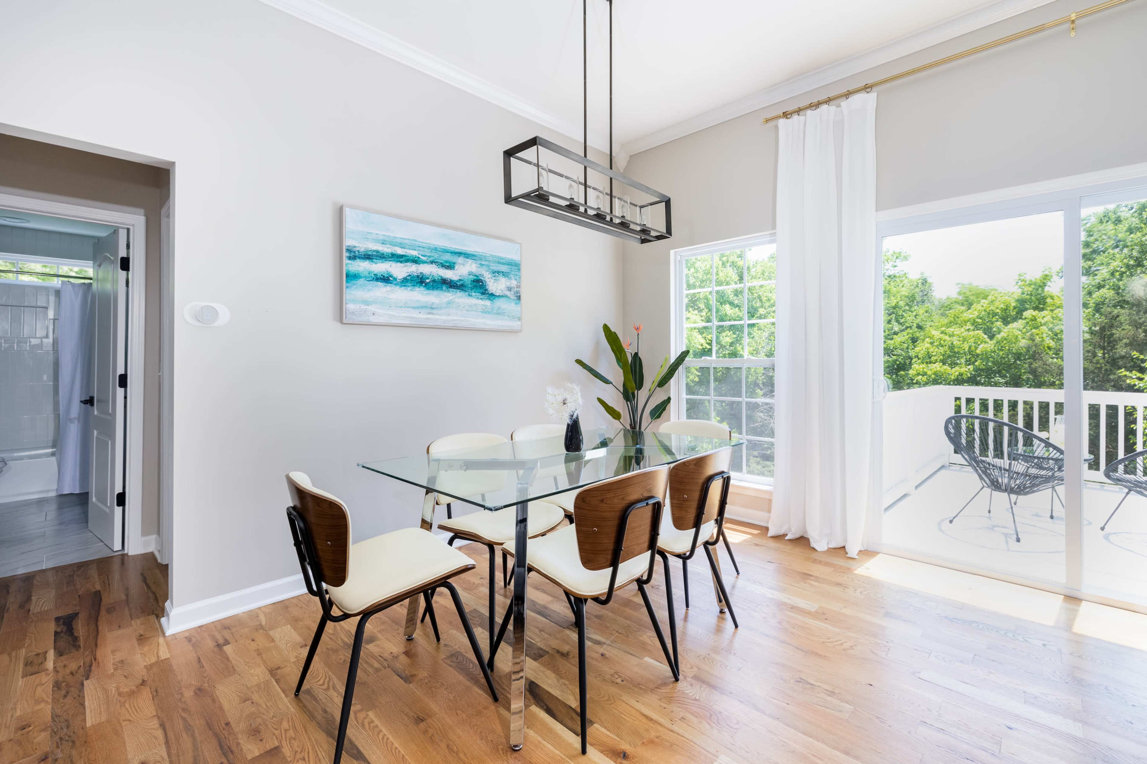 A modern dining area features a glass table with six chairs, a framed ocean painting on the wall, and large windows providing natural light and a view of greenery.
