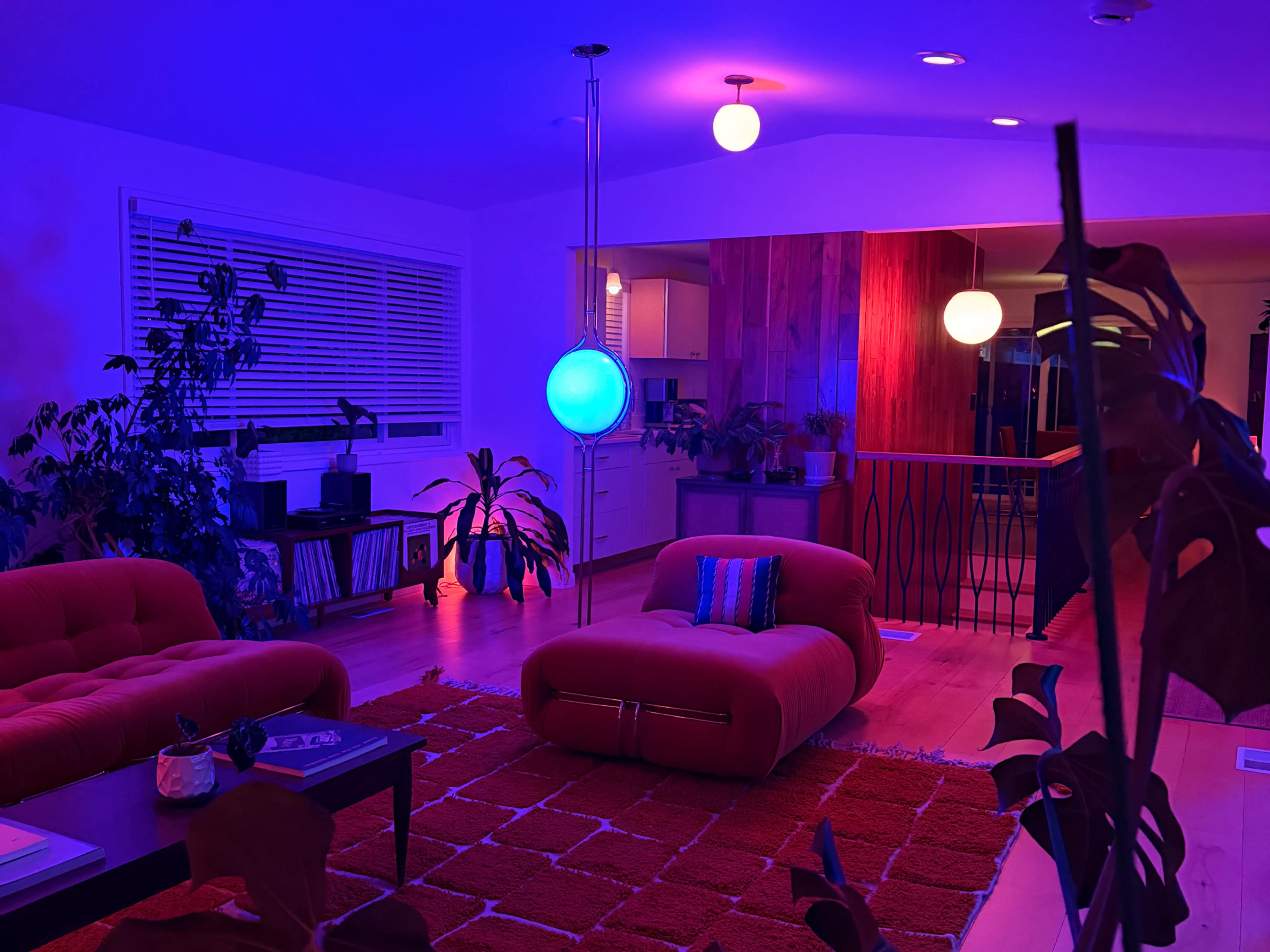 A brightly lit living room features red couches, a round rug, and colorful lighting from pendant fixtures.