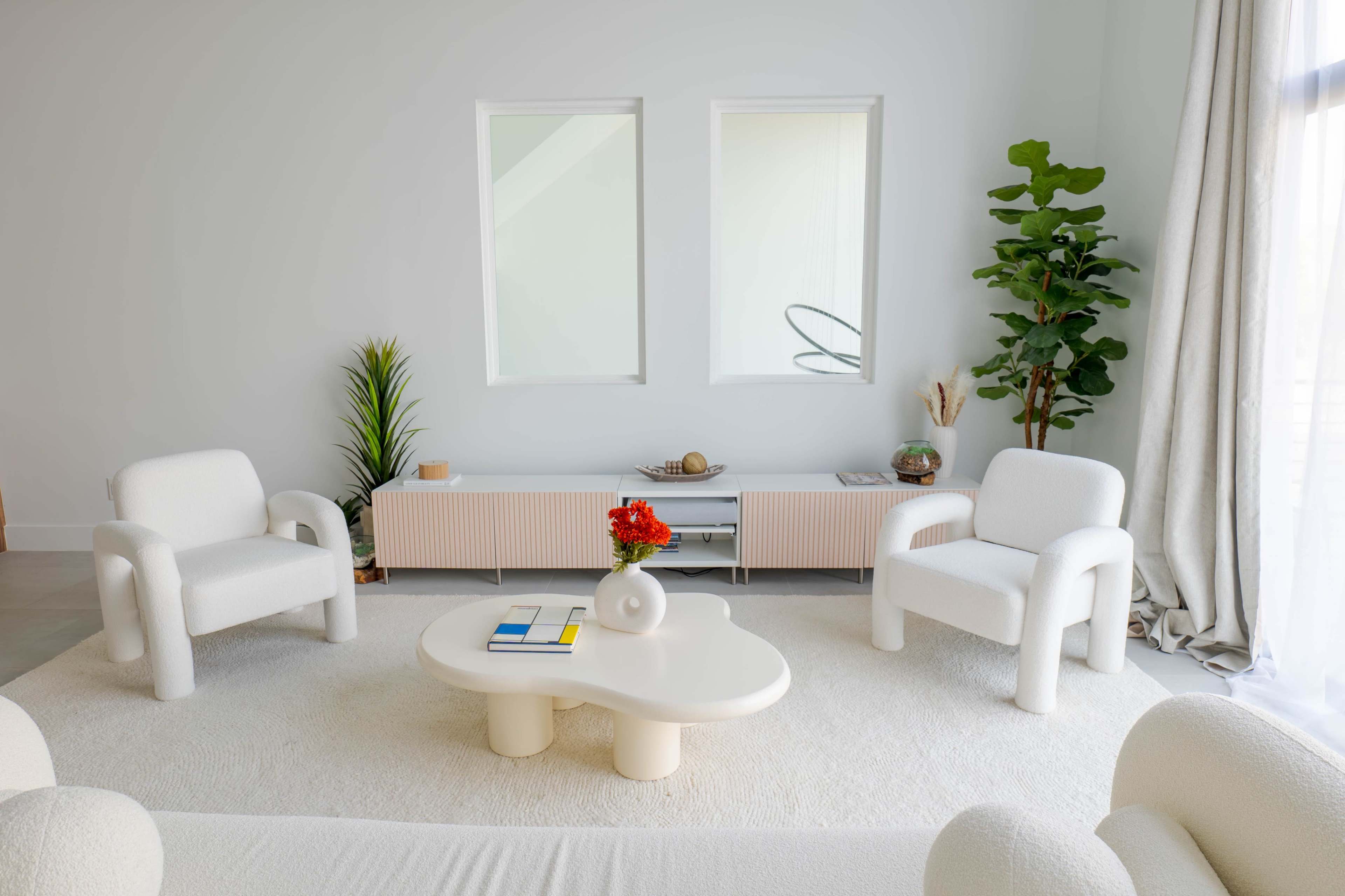 A minimalist living room features two chairs, a round coffee table, and a low sideboard, with large windows allowing natural light to enter.