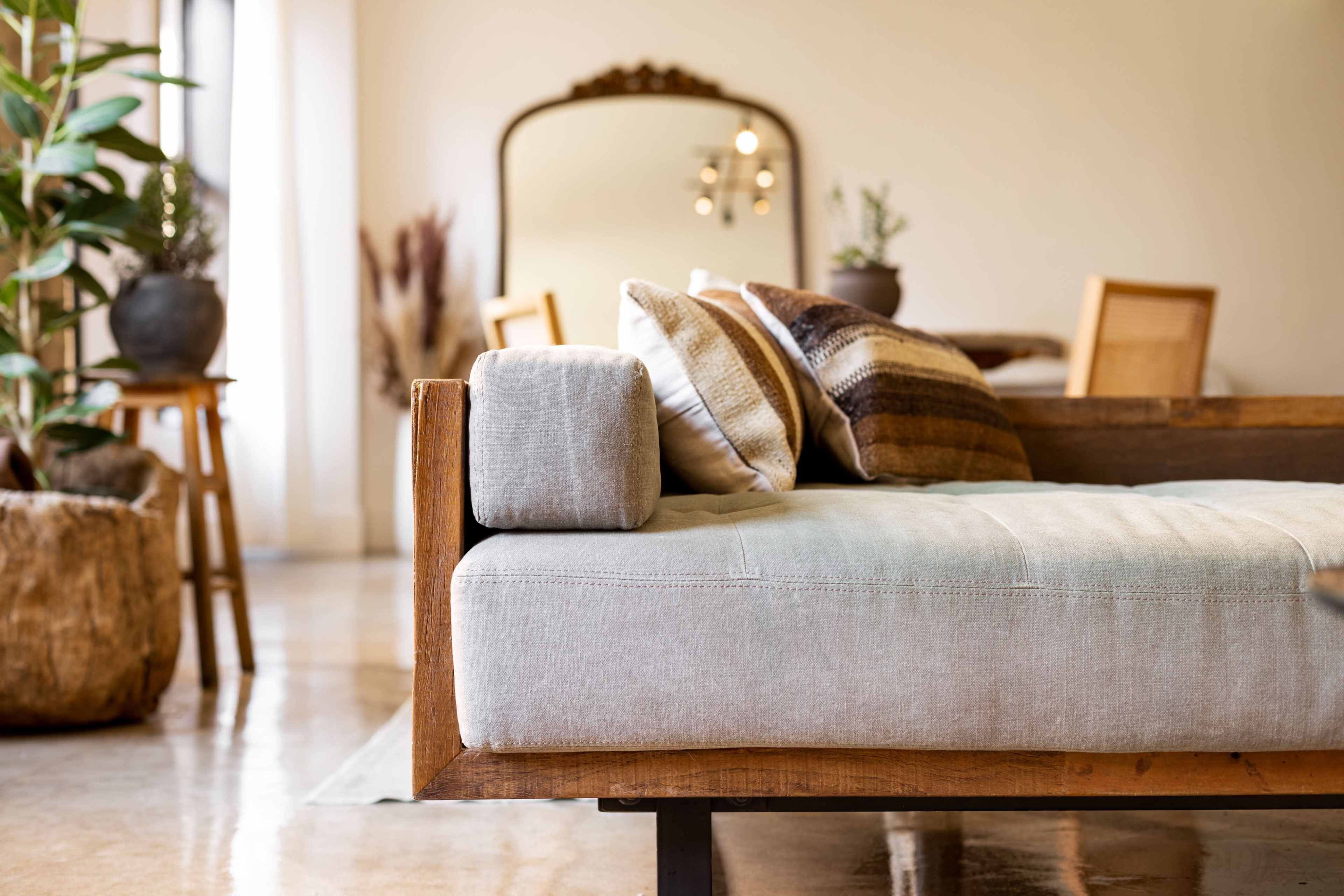 A wooden sofa with a light-colored fabric cushion and decorative pillows is positioned in a bright, stylish living space featuring plants and a mirrored wall.