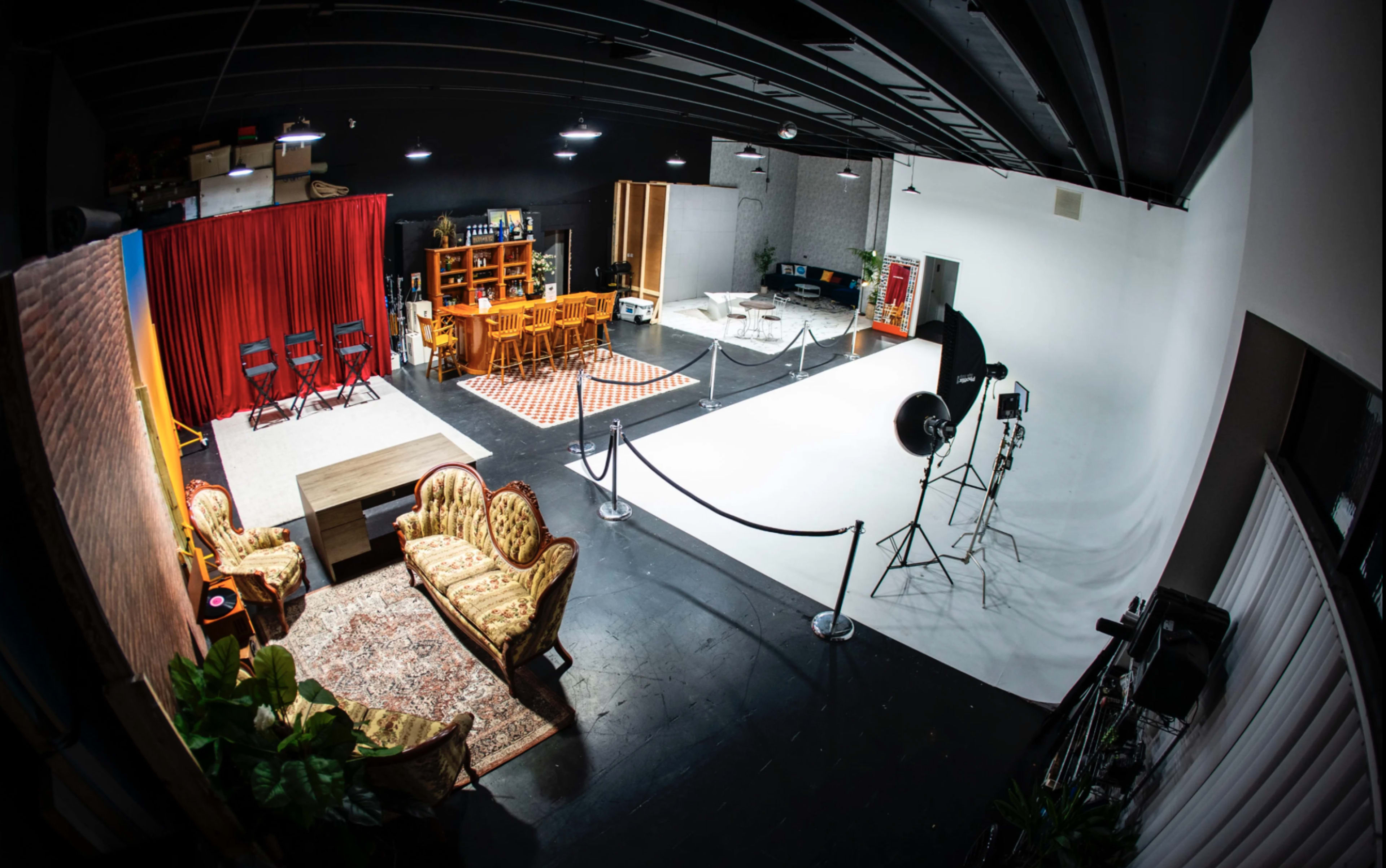 The image shows a large studio space divided into areas with a seating arrangement, dining set, and a white backdrop for filming.