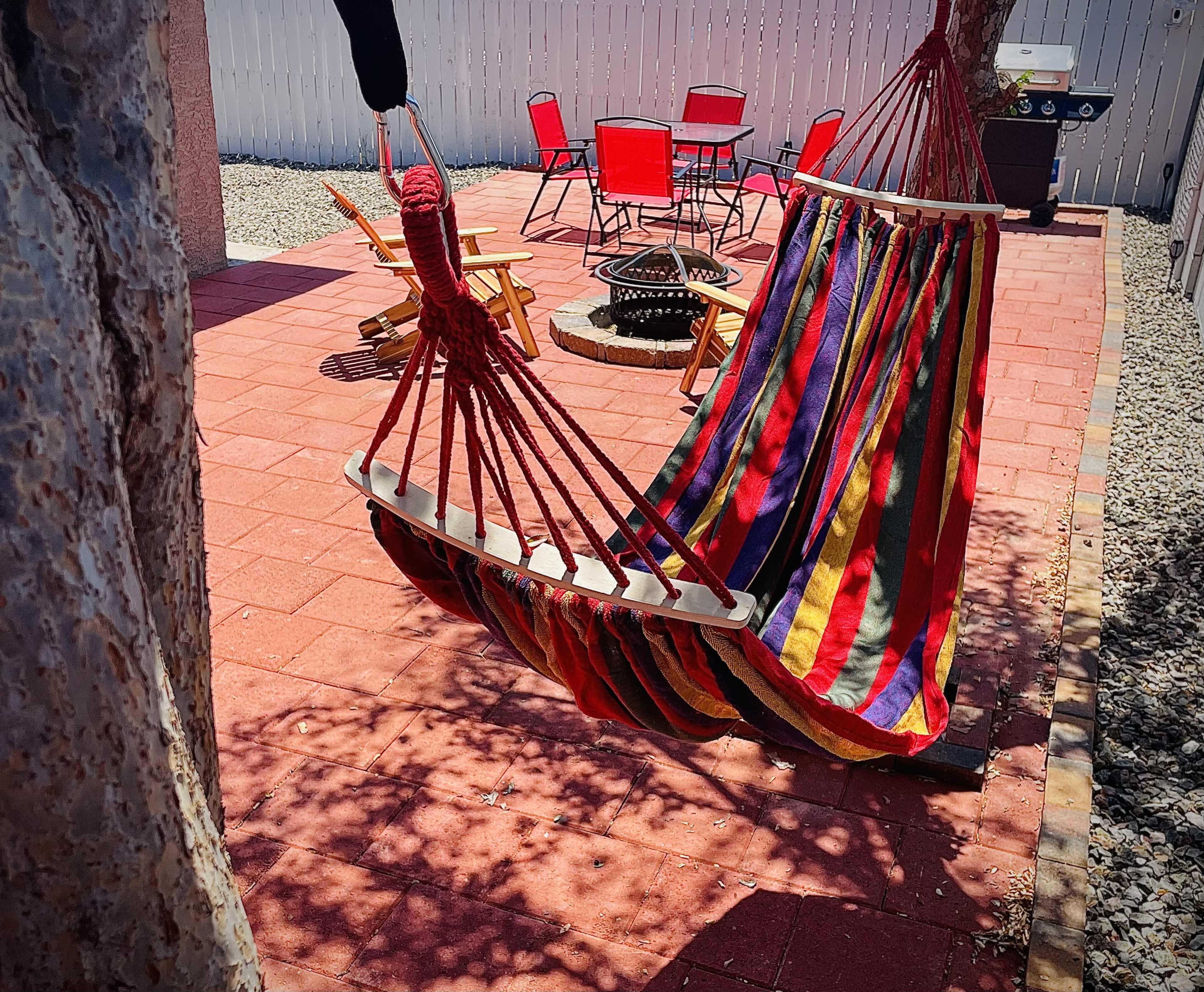 A colorful hammock hangs between two trees in a patio area with red chairs and a fire pit.