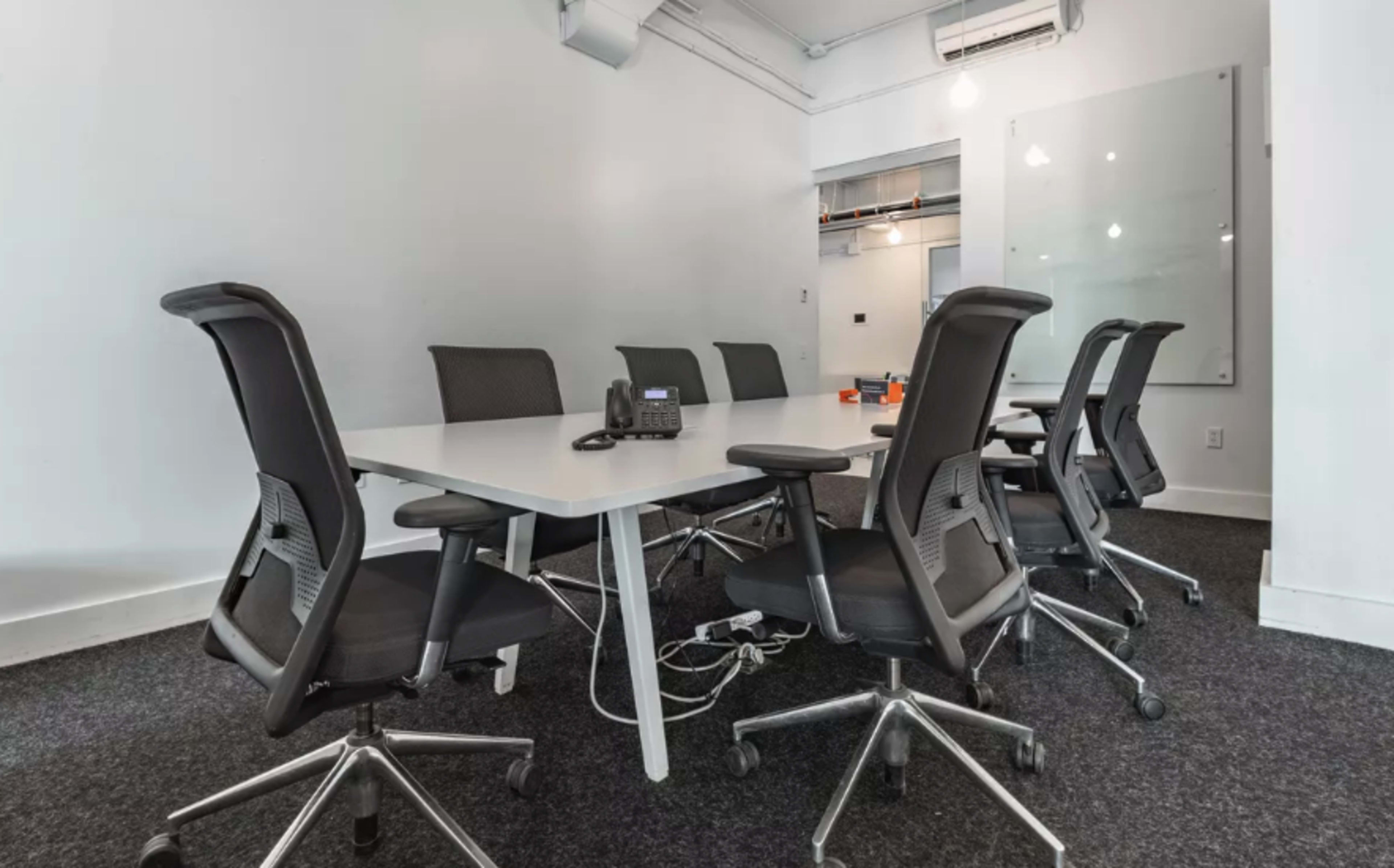 A modern conference room with a white table surrounded by six black office chairs and a phone on the table.