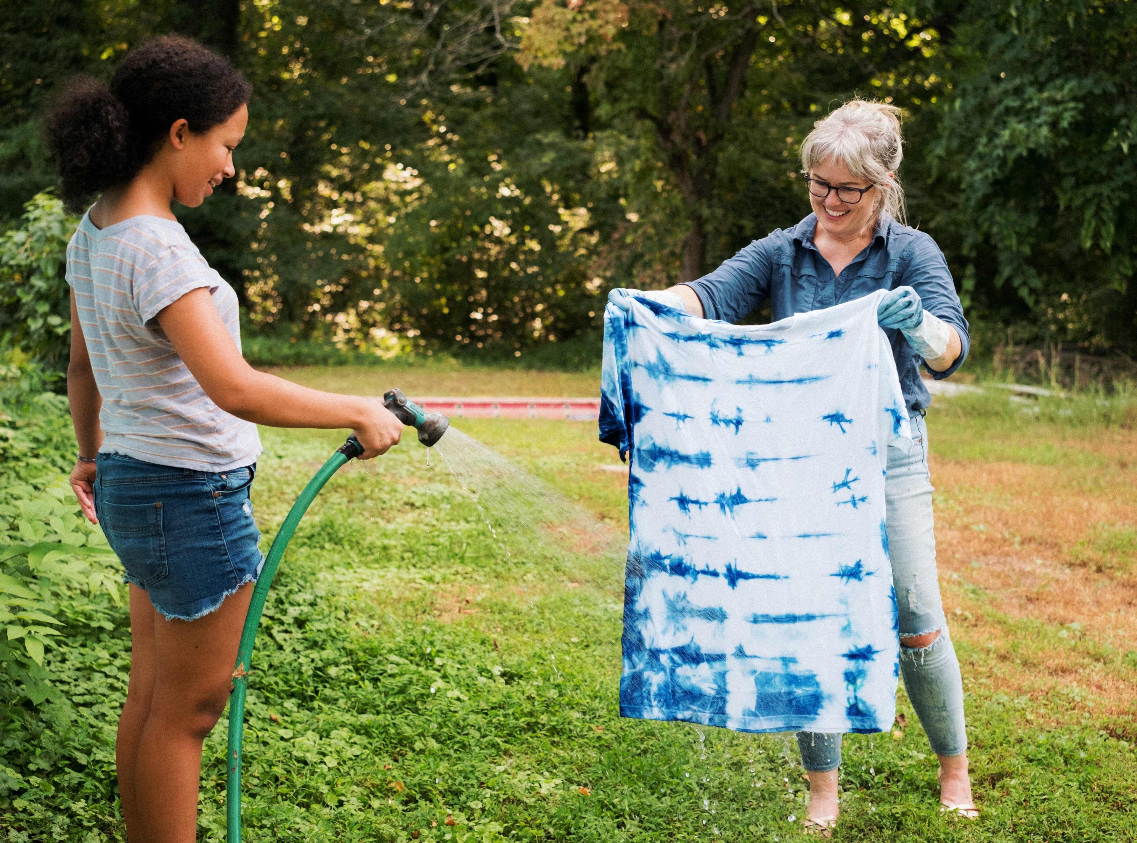 A woman holds a tie-dyed shirt while another sprays it with a hose in a grassy outdoor setting.